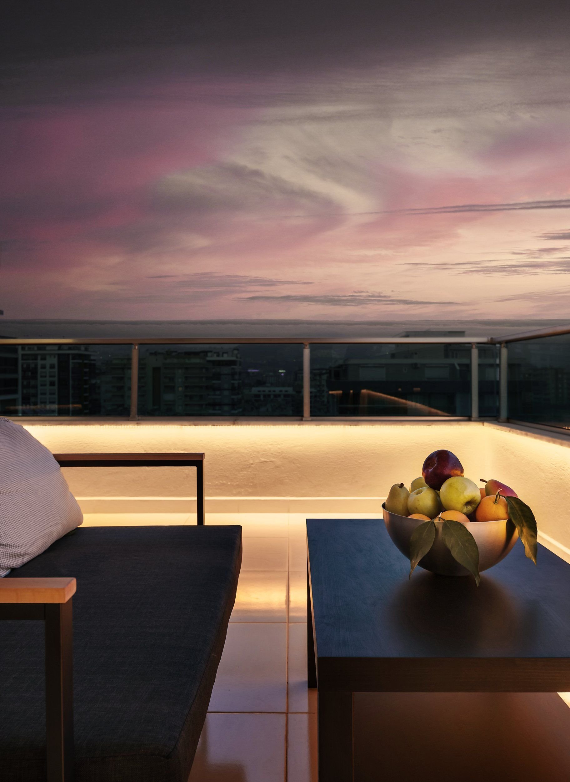Dämmerungsblick vom Dach. Eine Obstschale auf einem Tisch, eine Couch und eine Skyline im Hintergrund unter rosa Himmel.