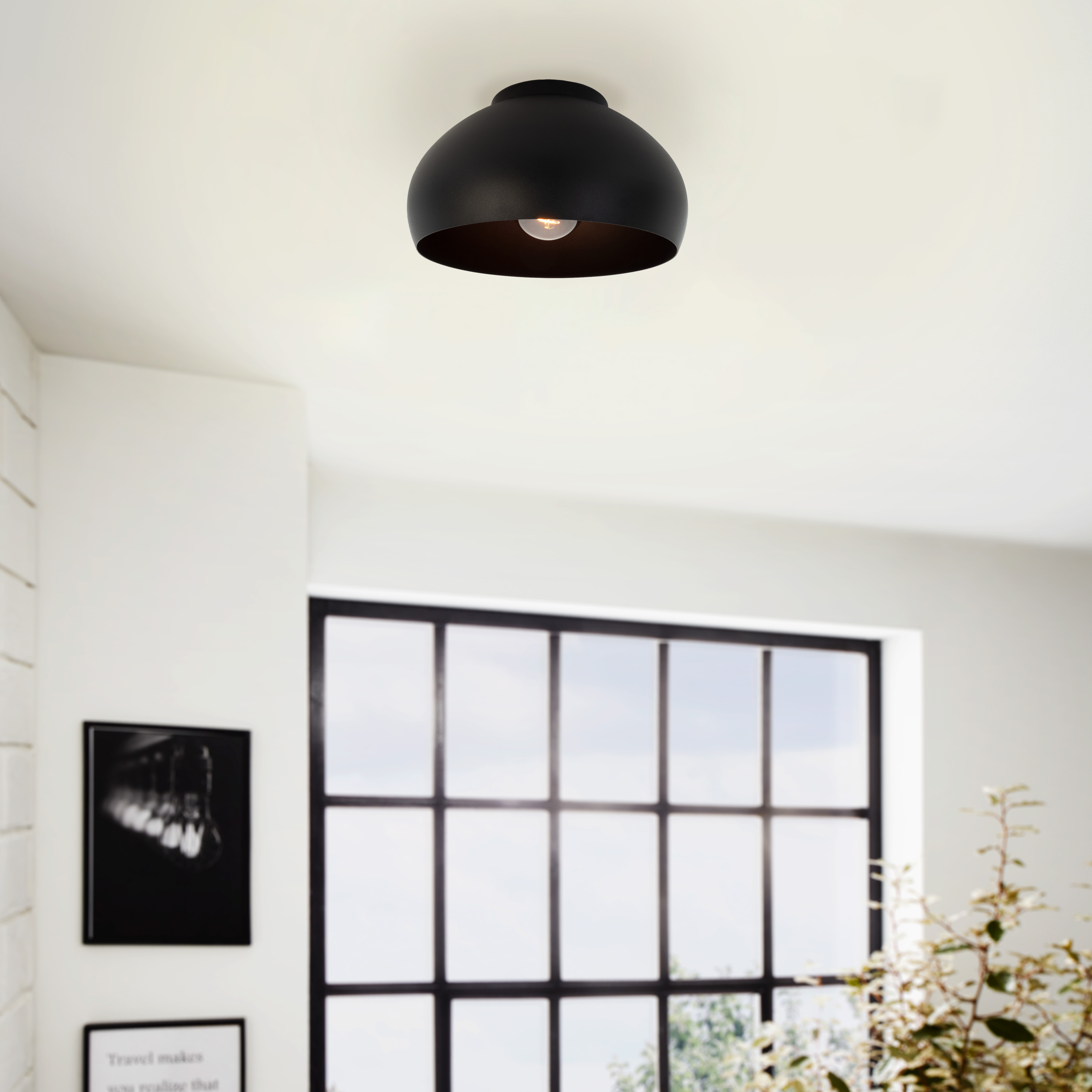 Una lampada da soffitto nera con una lampadina è centrata. Una finestra e delle piante sullo sfondo. Si vedono muri bianchi.