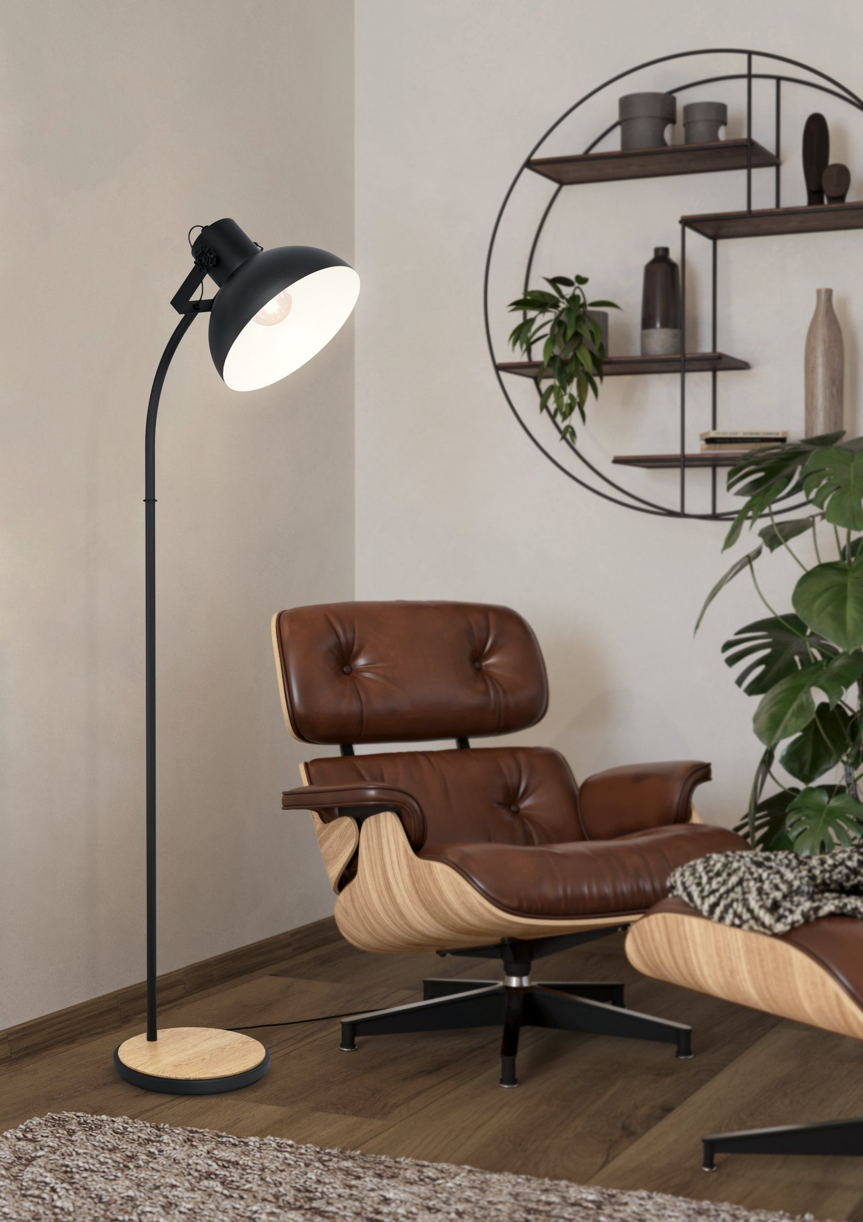 Une lampe noire éclaire un fauteuil et un pouf en cuir marron. Un mur avec des étagères et des plantes complète la scène.