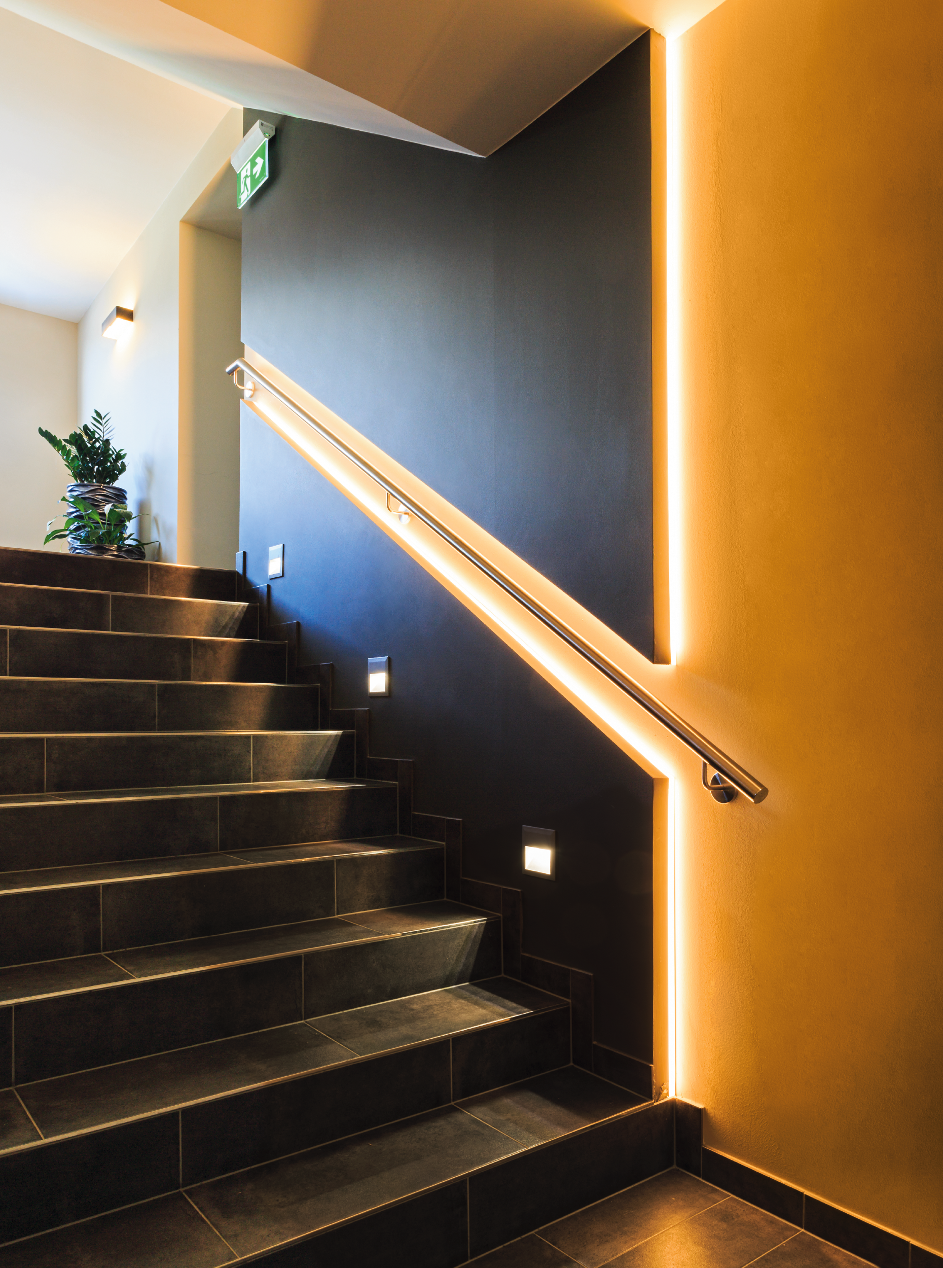 Treppe mit dunklen Fliesen, beleuchtet von einem Handlauf und Wandleuchten. Eine Pflanze steht auf dem Treppenabsatz. Notausgangsschild.