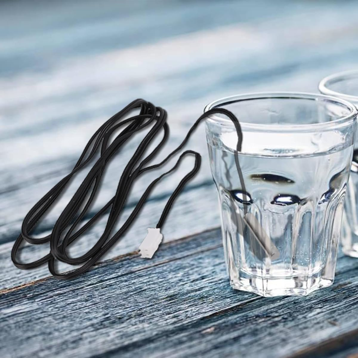 Ein schwarzes Kabel mit weißem Stecker ist mit einem Wasserglas verbunden. Das Glas steht auf einer Holzoberfläche.