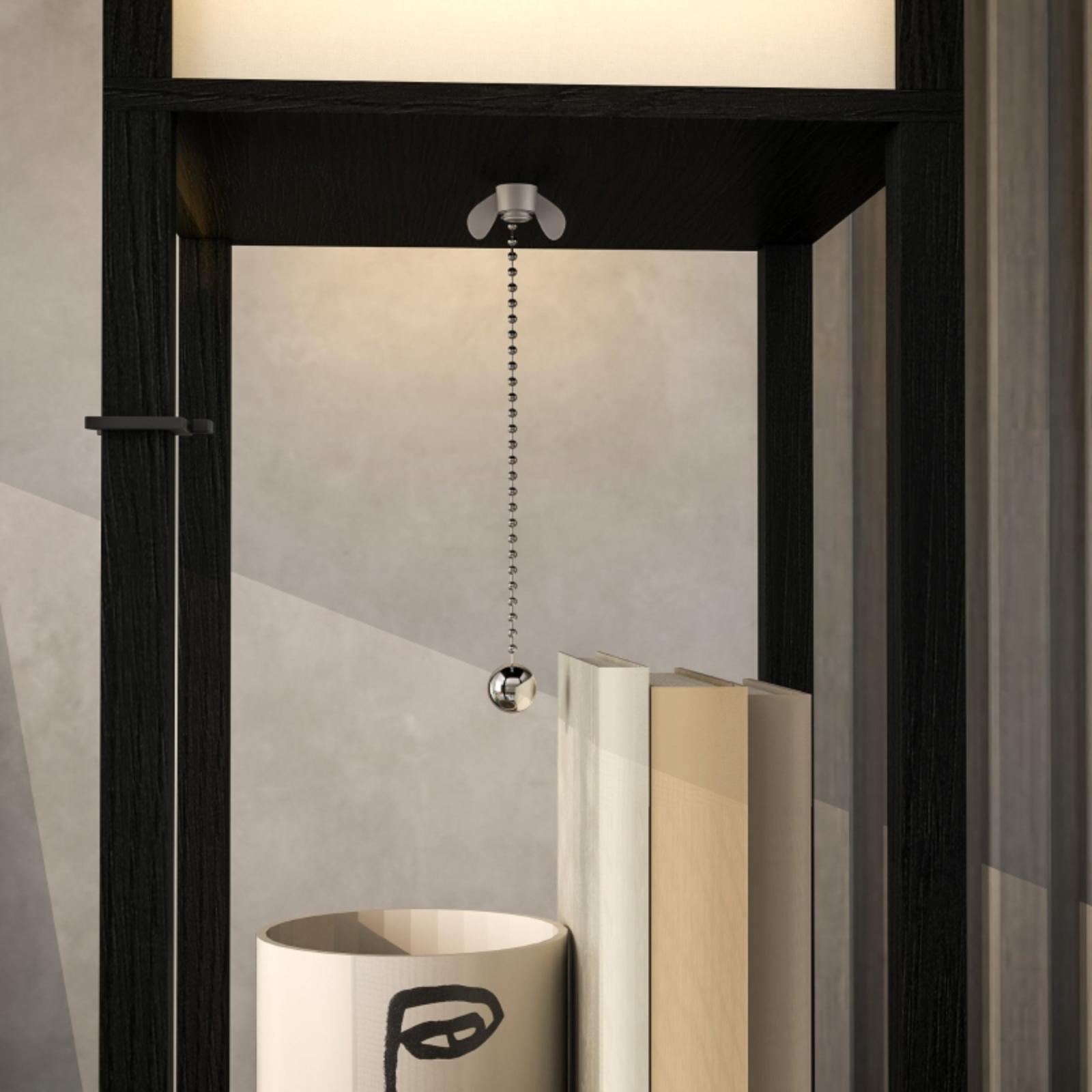 Ein schwarz gerahmtes Regal mit einer hängenden Kette. Darunter ein zylindrischer Behälter mit Gesichtsdesign und Bücher.