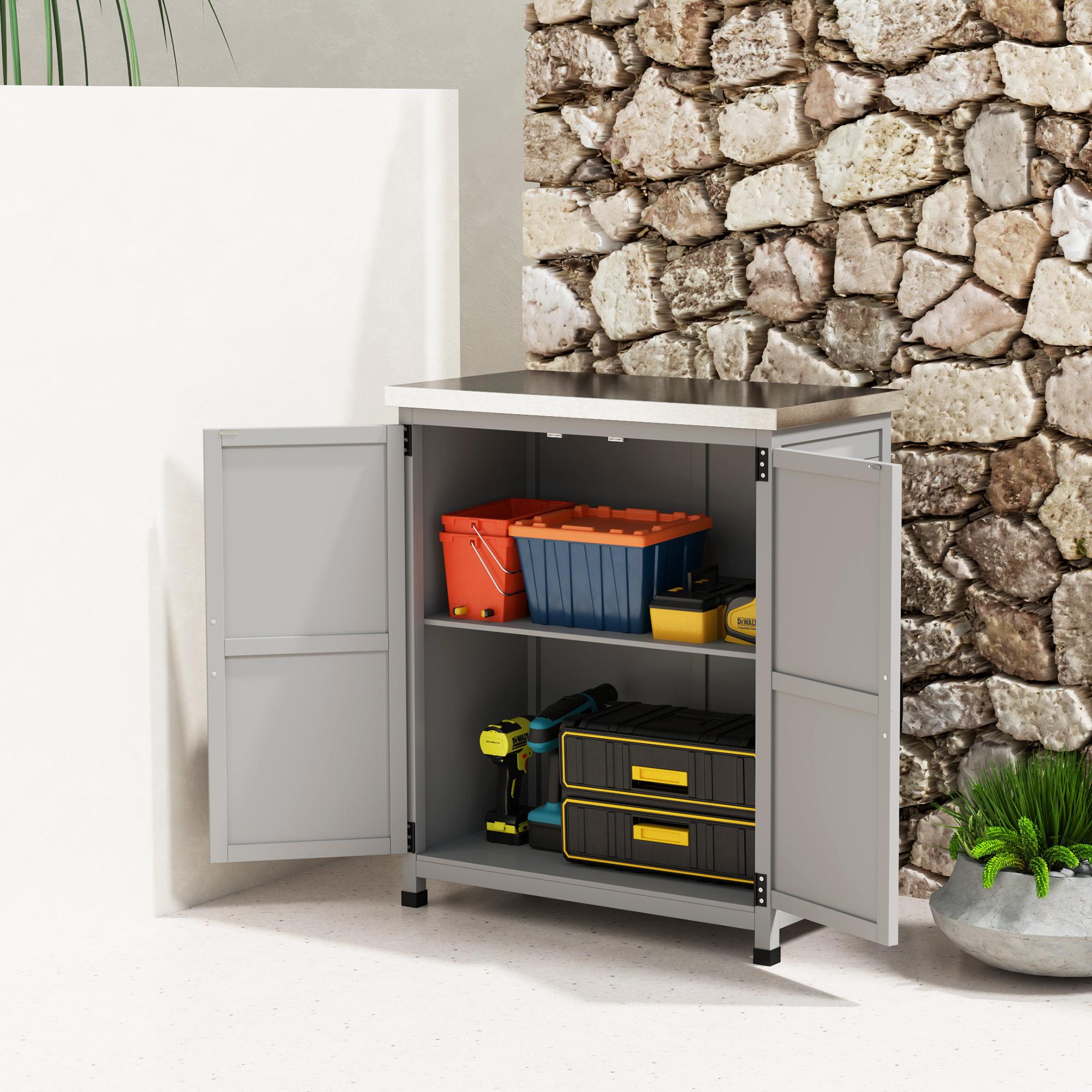 Ein grauer Schrank mit offenen Türen. Im Inneren befinden sich Werkzeuge und Aufbewahrungsboxen. Im Hintergrund eine Steinmauer und eine Pflanze.