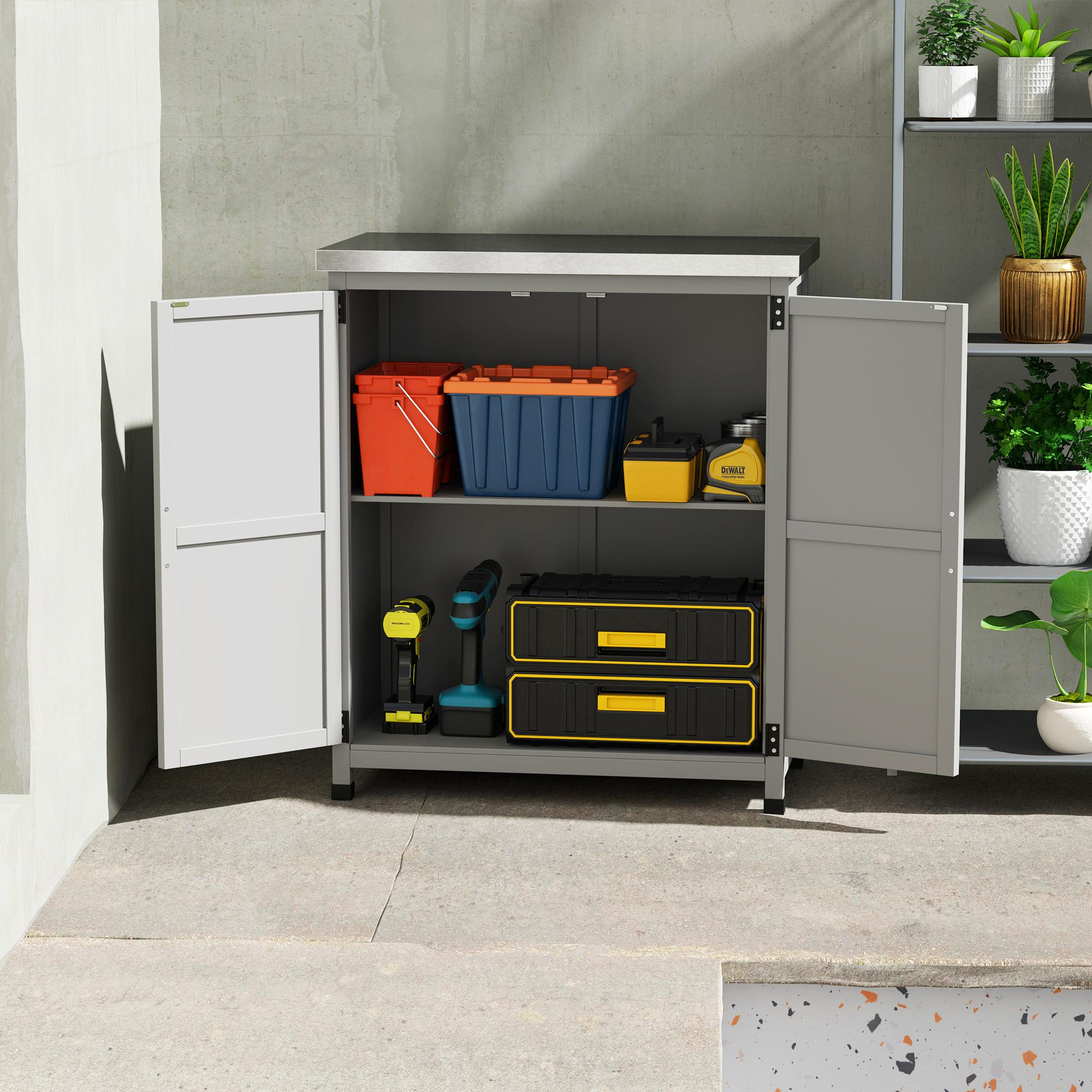 Ein grauer Schrank mit offenen Türen, der Werkzeuge und Behälter enthält. Auf der rechten Seite befindet sich ein Pflanzenregal. Betonboden und -wand.