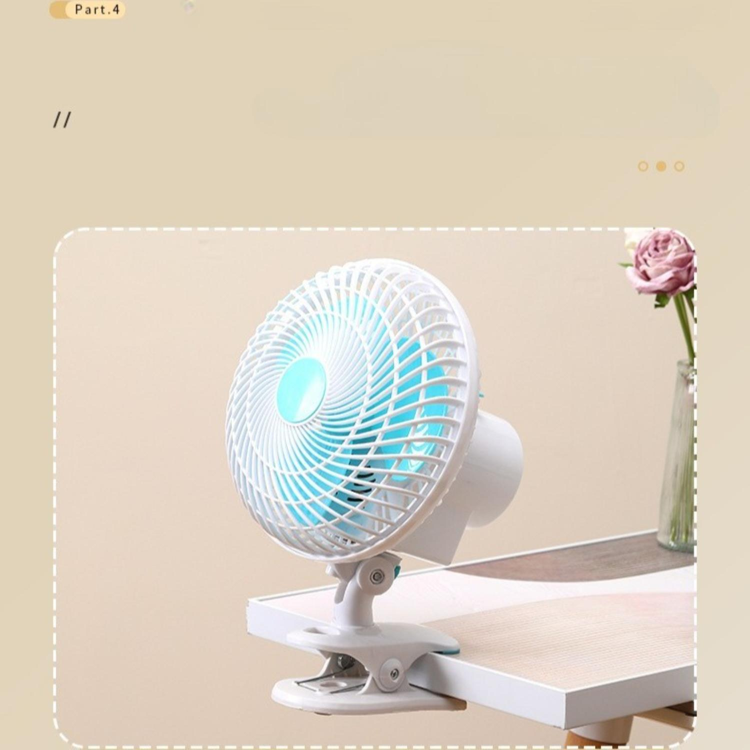 Ein kleiner weißer Ventilator mit blauen Flügeln steht auf einem Tisch. Eine Vase mit einer rosa Blume steht auf dem Tisch. Der Hintergrund ist beige.