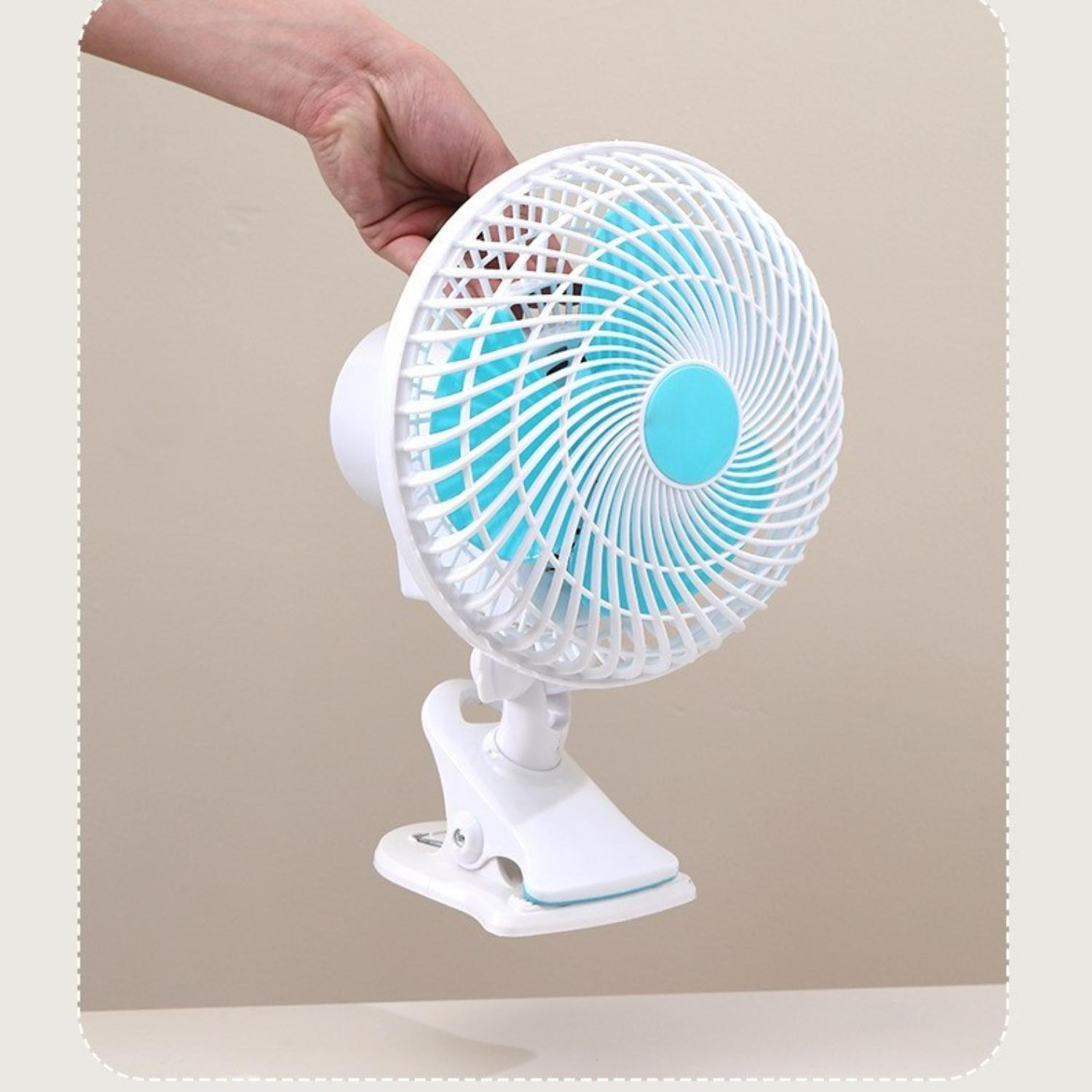 Ein weiß-blauer Clip-On-Ventilator wird von einer Hand gehalten. Der Lüfter hat einen weißen Käfig und blaue Flügel. Der Hintergrund ist hellbeige.