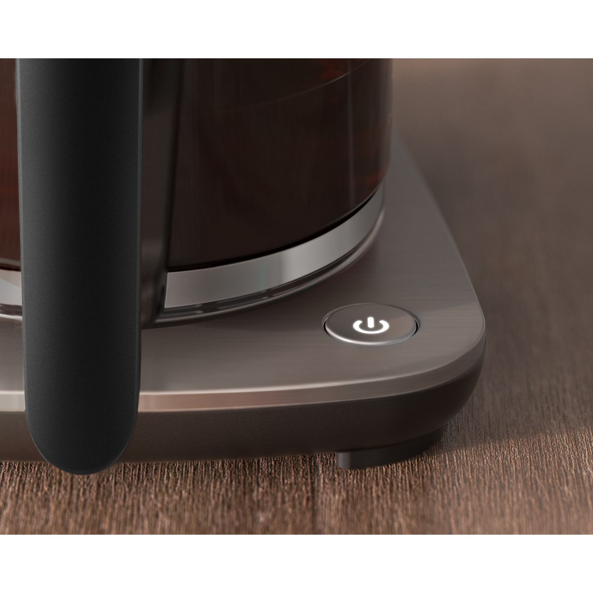 Zbliżenie na podstawę ekspresu do kawy z przyciskiem zasilania. Szklana karafka z ciemnym płynem na wierzchu. Drewniana powierzchnia w tle.