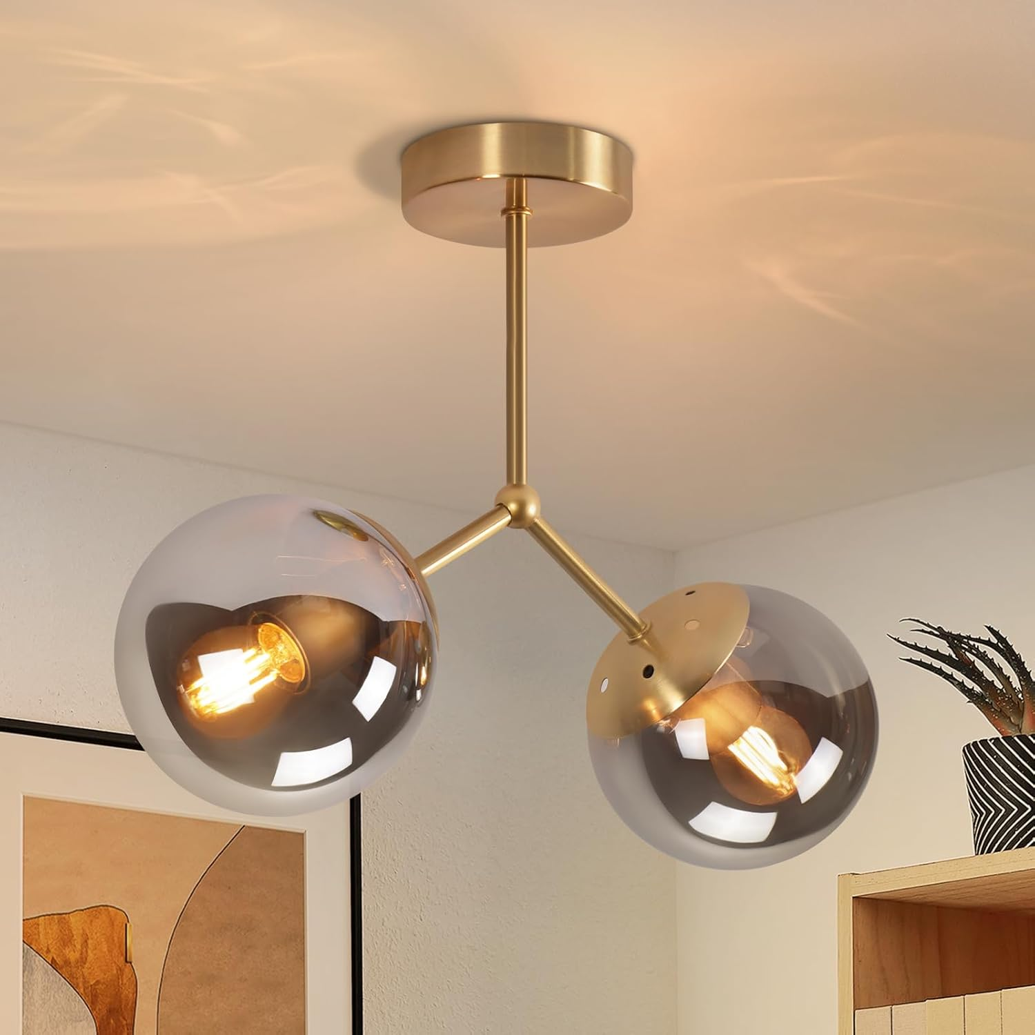 Złota lampa sufitowa z dwiema szklanymi kulami i centralną łodygą. Żarówki świecą, tło to biały sufit.