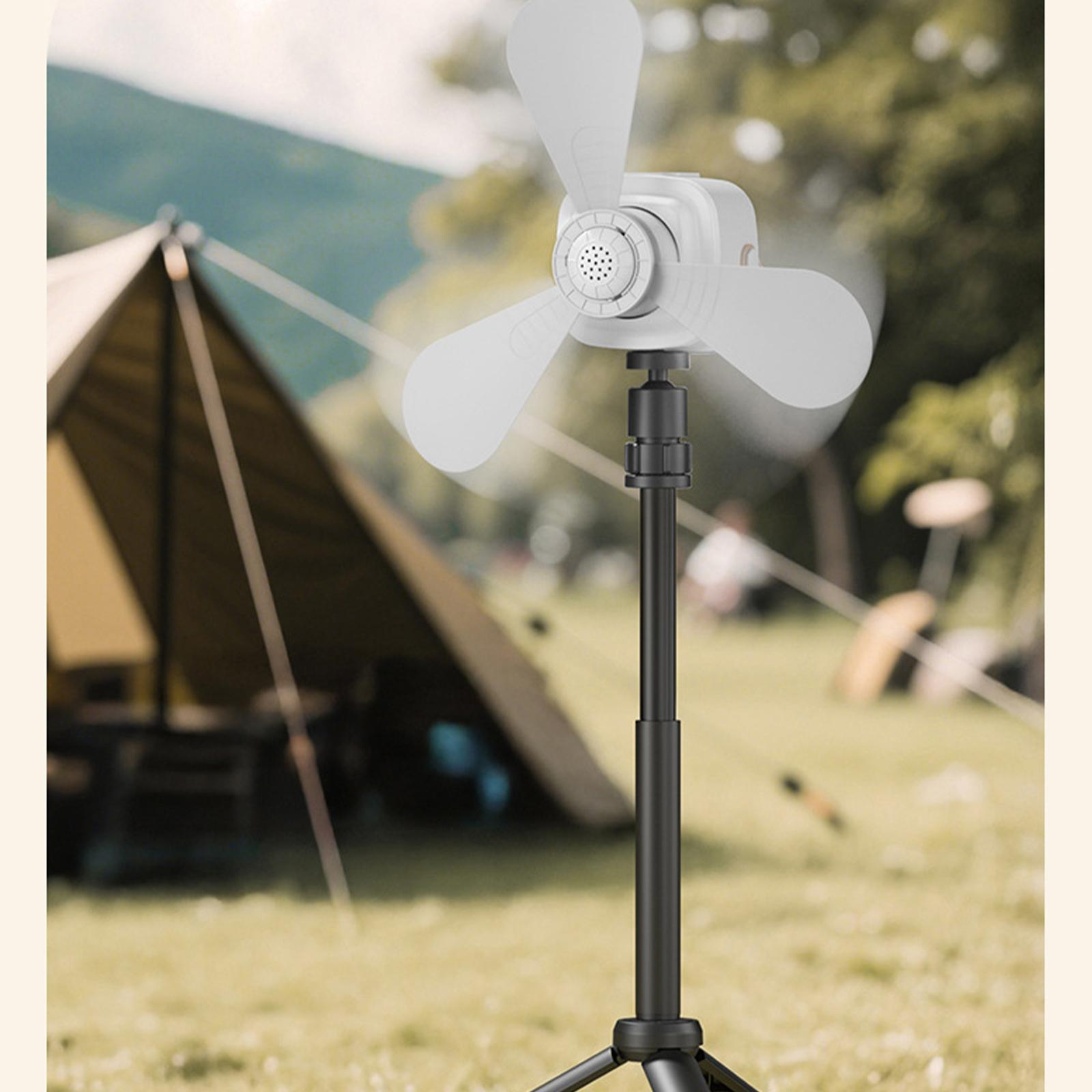 Ein weißer Ventilator mit drei Flügeln steht auf einem schwarzen Ständer. Im Hintergrund befindet sich ein Zelt auf einer grünen Wiese.