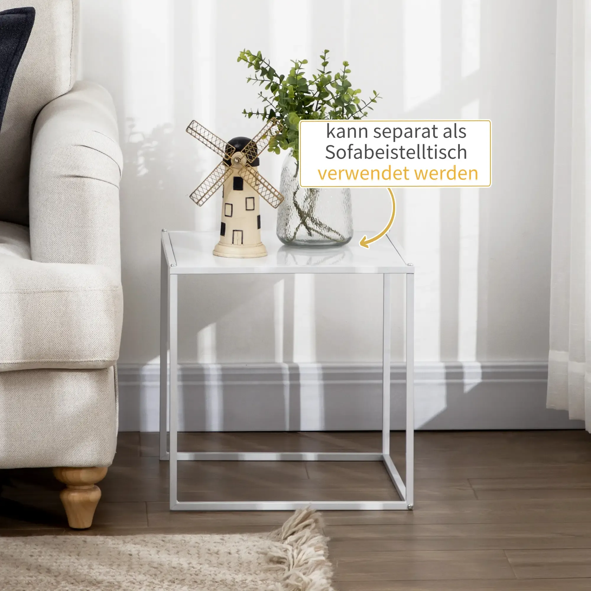 Weißer Beistelltisch mit Windmühlendekor und grüner Pflanze. Sofa im Hintergrund. Text-Overlay: kann separat als Beistelltisch verwendet werden.