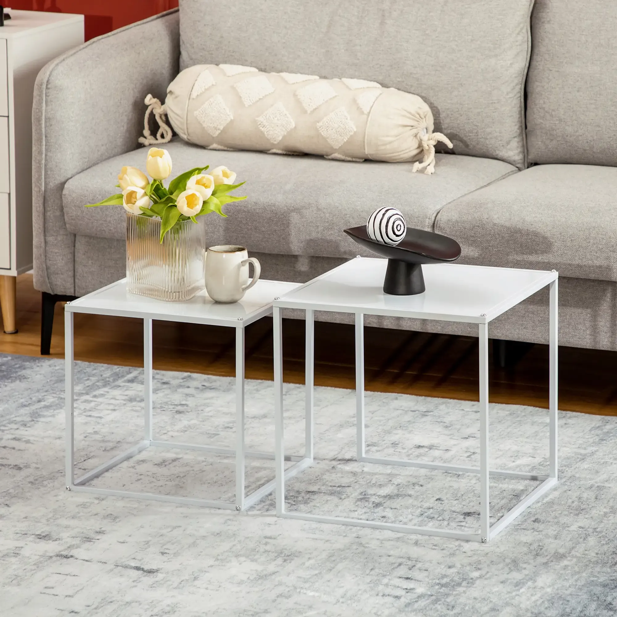 Zwei weiße Tische mit Objekten: Blumen, Becher und Deko. Graues Sofa dahinter. Grauer Teppich.