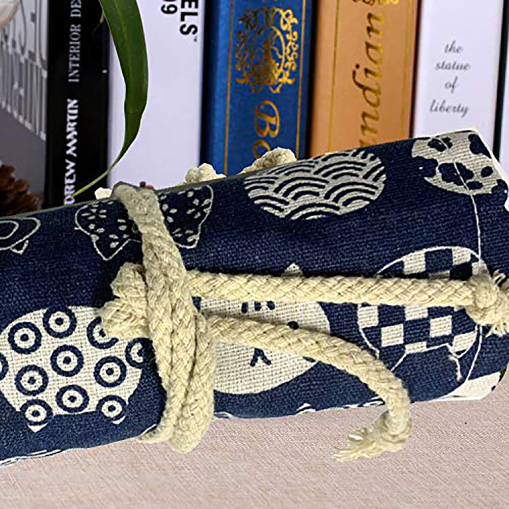 Ein gerolltes blaues Federmäppchen mit Muster, das mit beigem Seil zusammengebunden ist. Bücher sind im Hintergrund.