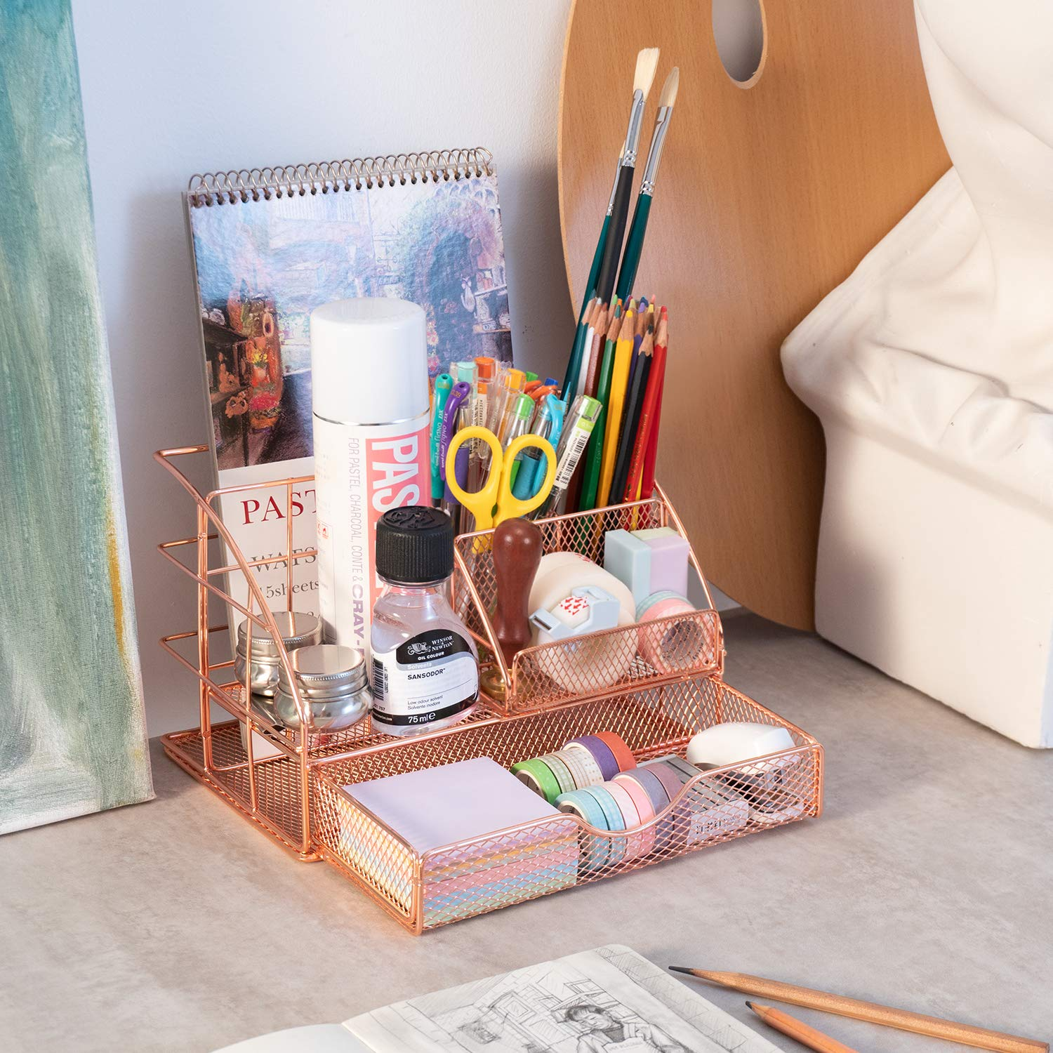 Roségoldfarbener Schreibtisch-Organizer mit Kunstutensilien: Bleistifte, Pinsel, Klebeband, Schere und Notizblöcke. Auch eine Palette und eine Zeichnung sind sichtbar.