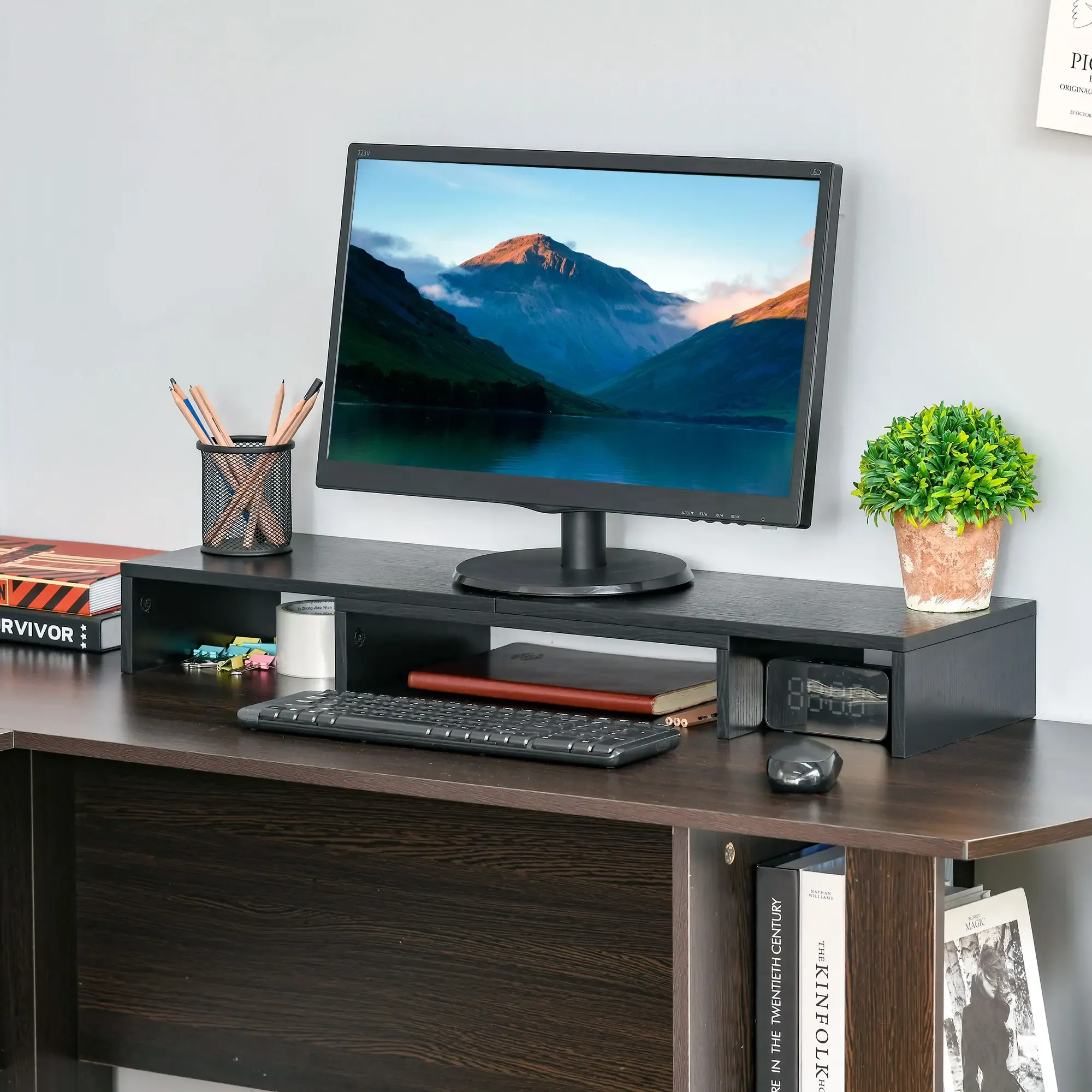 Schreibtisch mit Monitor, der eine Berglandschaft zeigt. Enthält Tastatur, Bleistifte, eine Pflanze, Bücher und eine Uhr.