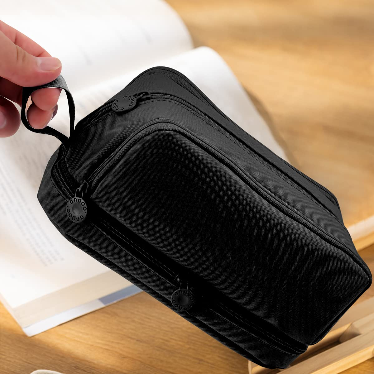 Schwarze rechteckige Tasche, die von einer Hand gehalten wird. Die Tasche befindet sich auf einer Holzoberfläche mit einem offenen Buch im Hintergrund.