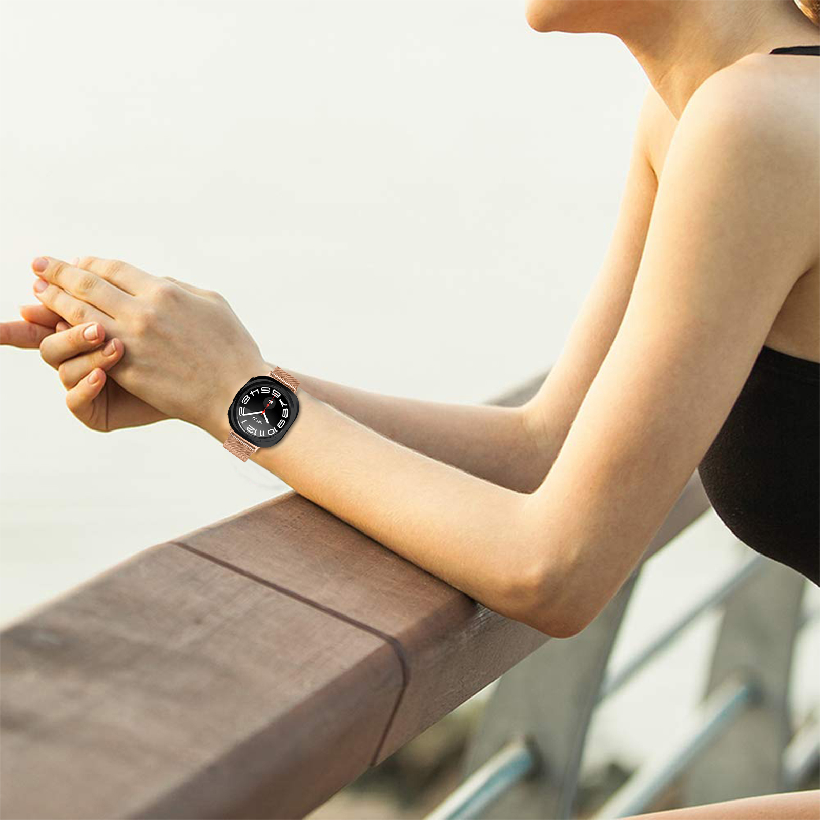 Eine Frau, die eine Smartwatch am Handgelenk trägt und sich auf ein Holzgeländer stützt. Die Uhr hat ein schwarzes Zifferblatt und ein roségoldenes Armband.