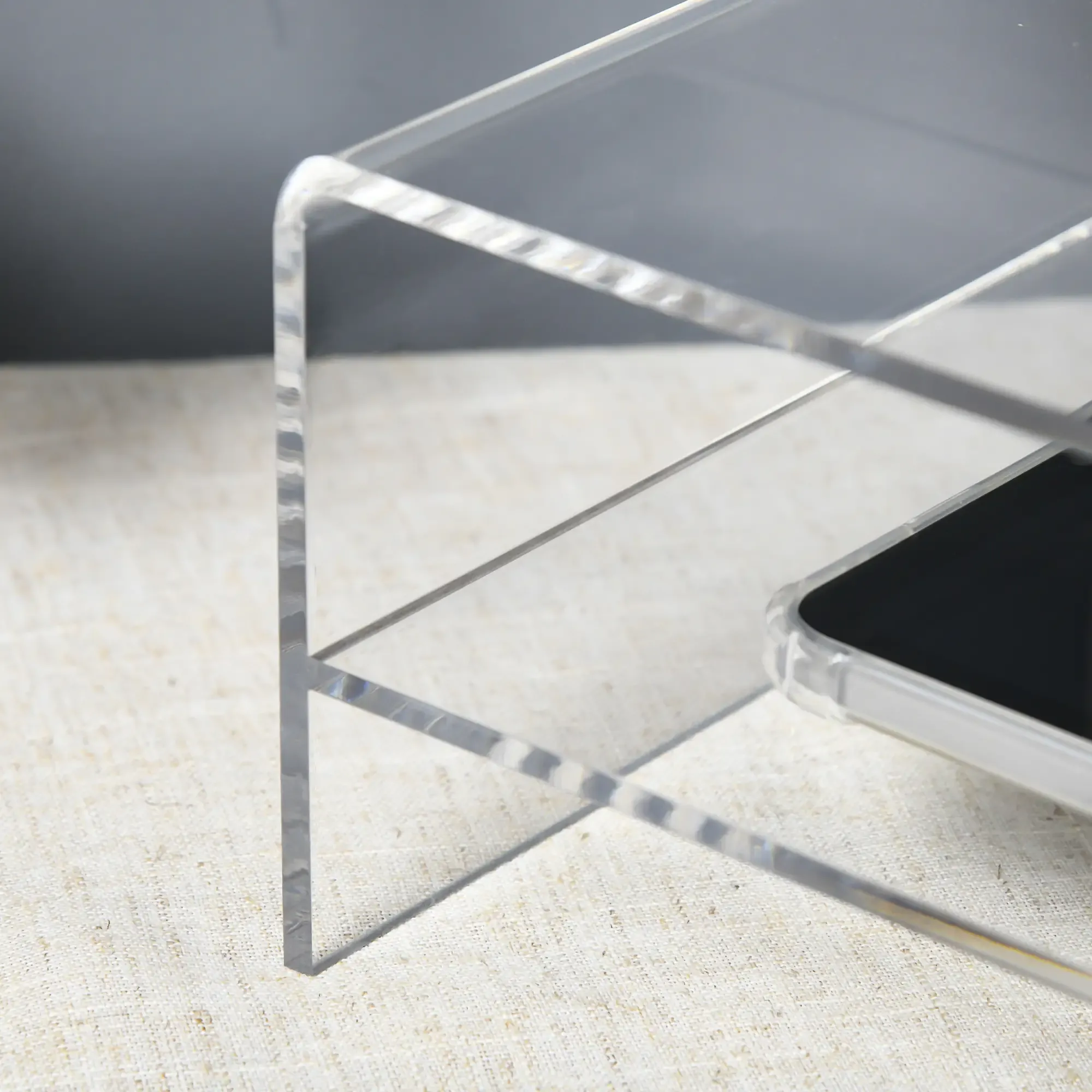 Klarer Acrylständer mit einem Telefon darauf. Der Ständer steht auf einer strukturierten Oberfläche mit grauem Hintergrund.