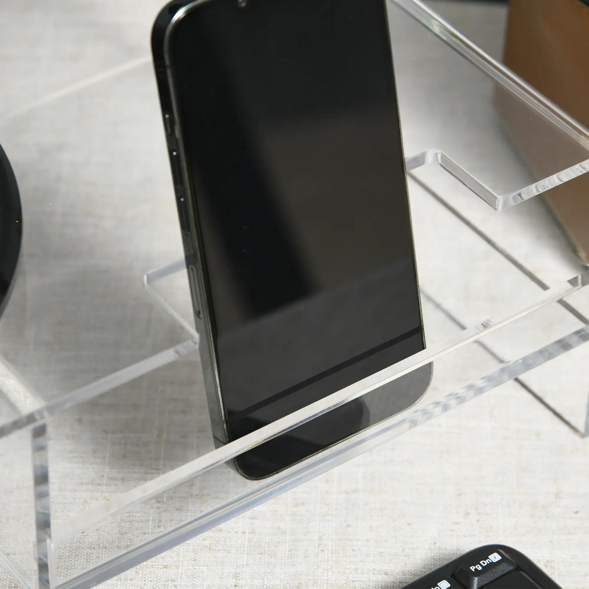 Ein schwarzes Smartphone liegt auf einem durchsichtigen Acrylständer. Tastatur in der Ecke. Der Hintergrund ist eine hellfarbige Oberfläche.
