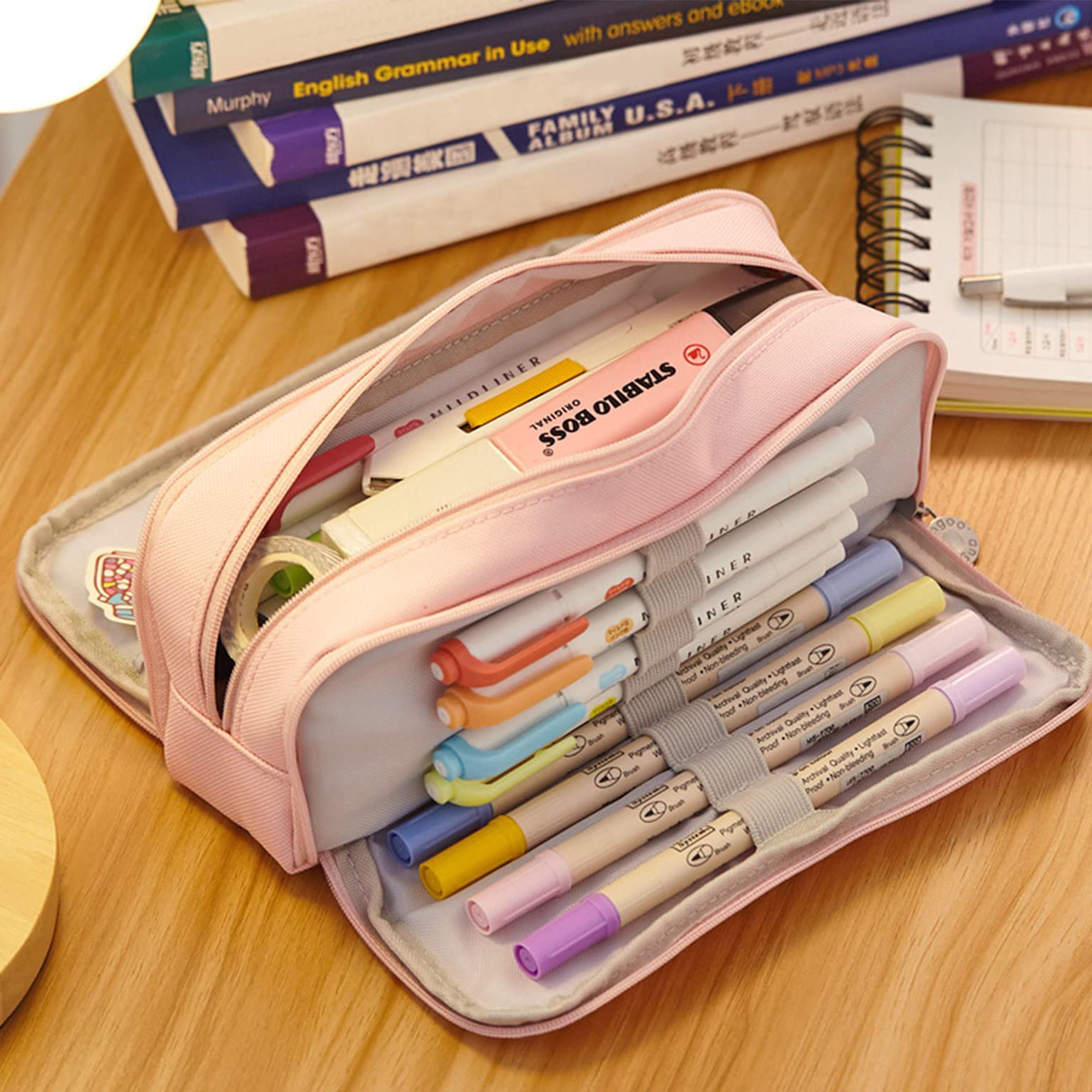Ein offenes, pinkfarbenes Federmäppchen mit Markern, Textmarkern und Klebeband. Bücher und ein Notizblock befinden sich im Hintergrund auf einem Holztisch.