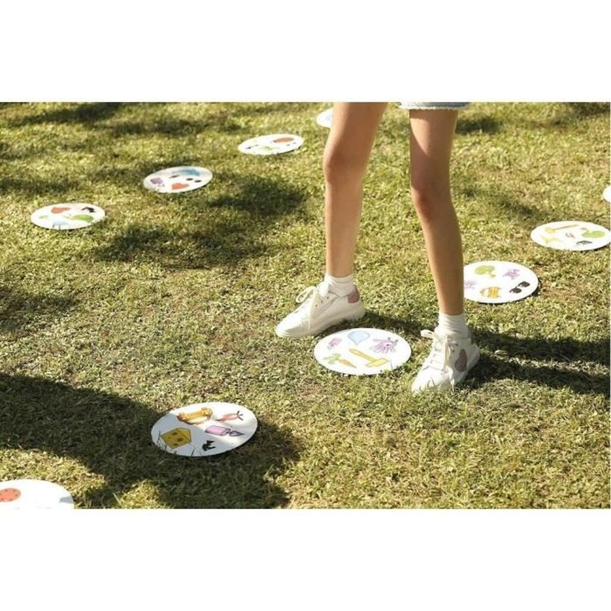 Die Beine einer Person und weiße Turnschuhe befinden sich auf einem Rasen mit runden Spielsteinen. Jedes Teil hat Zeichnungen. Das Gras ist grün.