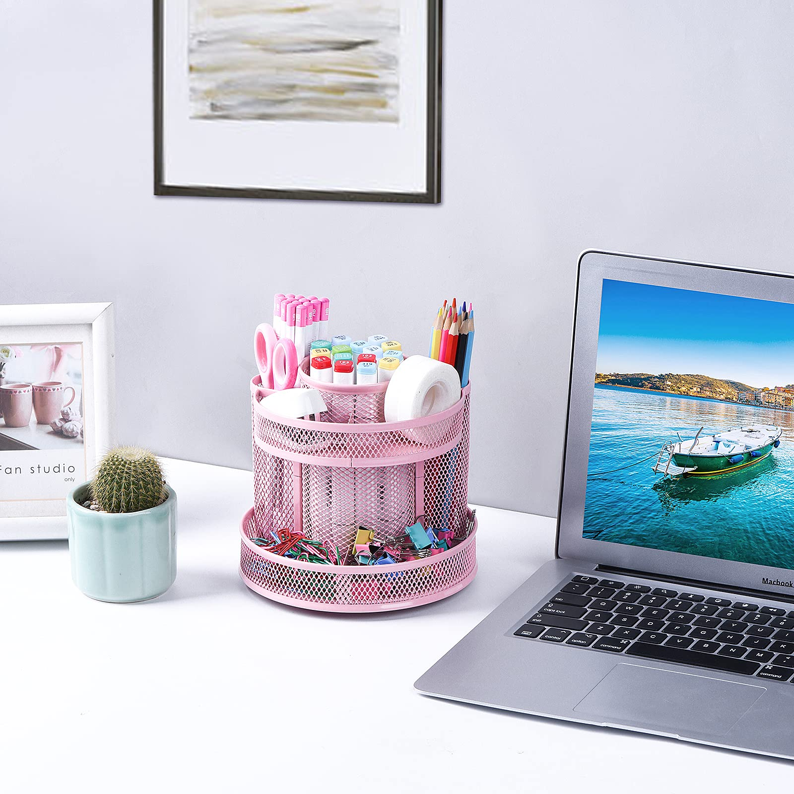 Eine Schreibtischszene mit einem rosa Stifthalter, einem Laptop mit einem Boot, einem Kaktus und einem gerahmten Bild.