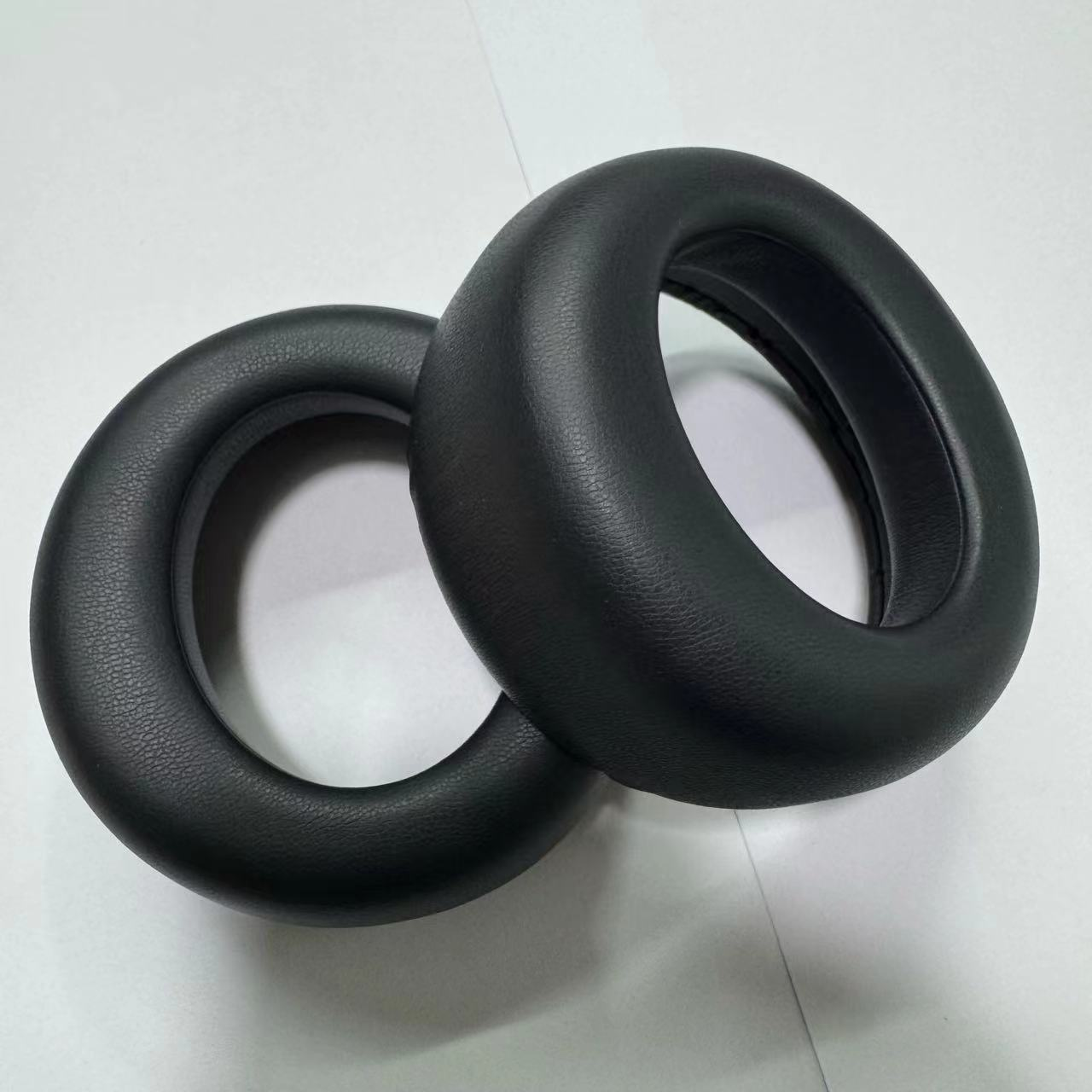 Due cuscinetti auricolari circolari neri per cuffie su una superficie bianca. Sono in materiale strutturato e hanno un foro al centro.