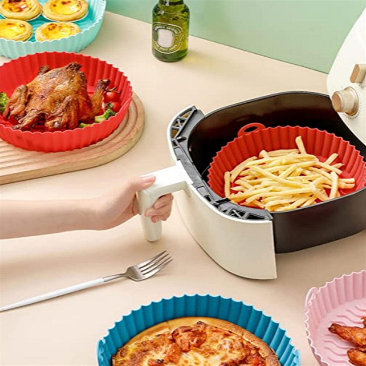 Ein Luftfritteuse mit Pommes, ein gebratenes Huhn, Pizza und Gebäck. Eine Hand hält die Luftfritteuse. Verschiedene bunte Tabletts sind vorhanden.
