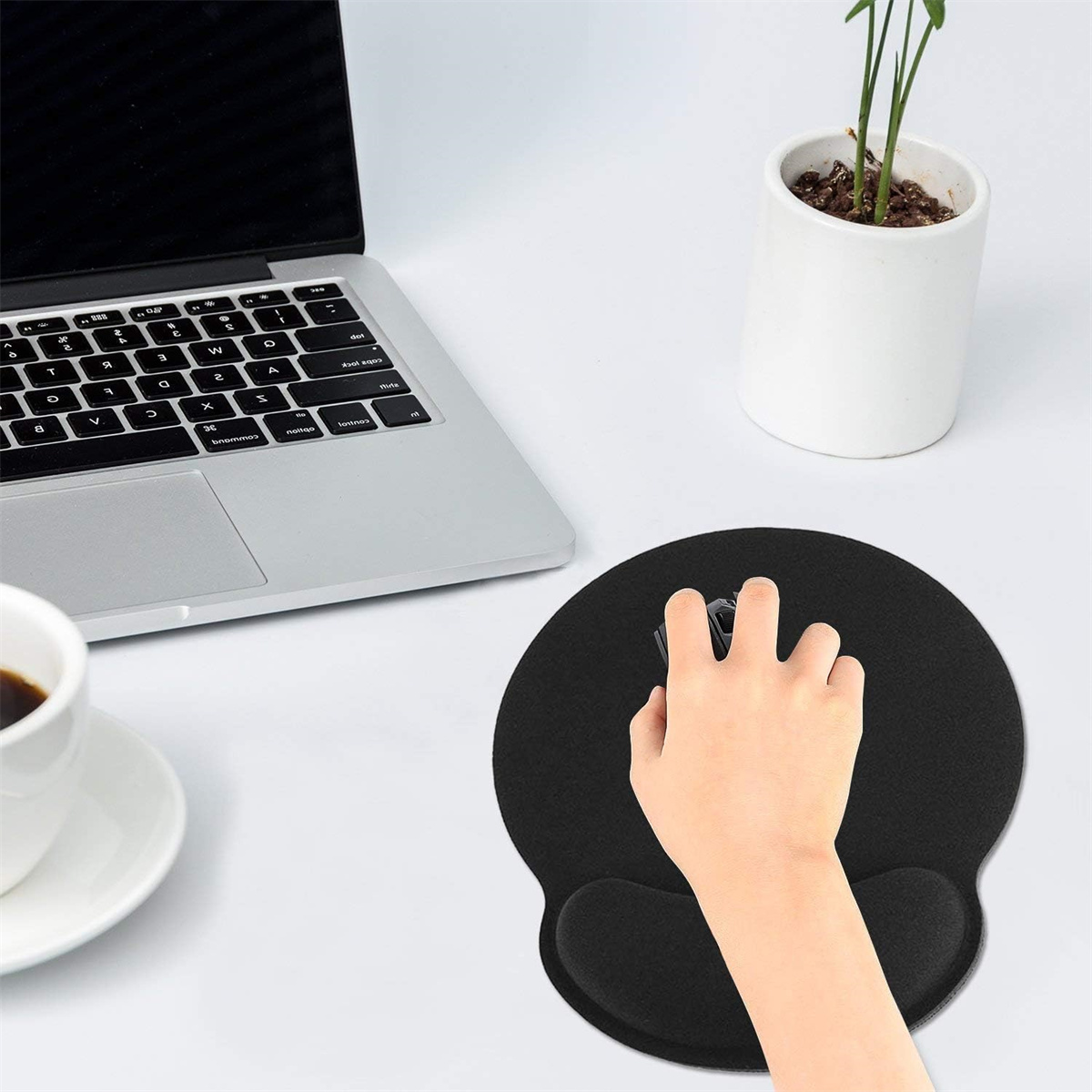 Eine Hand benutzt eine Maus auf einem schwarzen Mauspads. Ein Laptop, eine Kaffeetasse und eine Topfpflanze sind ebenfalls auf dem weißen Schreibtisch.