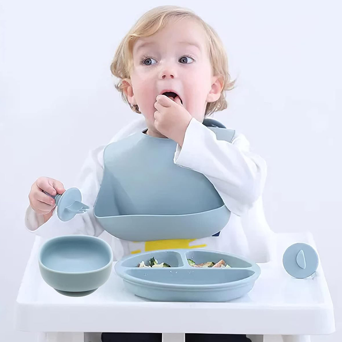 Ein Baby mit blonden Haaren isst. Es gibt eine blaue Latzhose und Geschirr. Das Baby sitzt in einem weißen Hochstuhl.