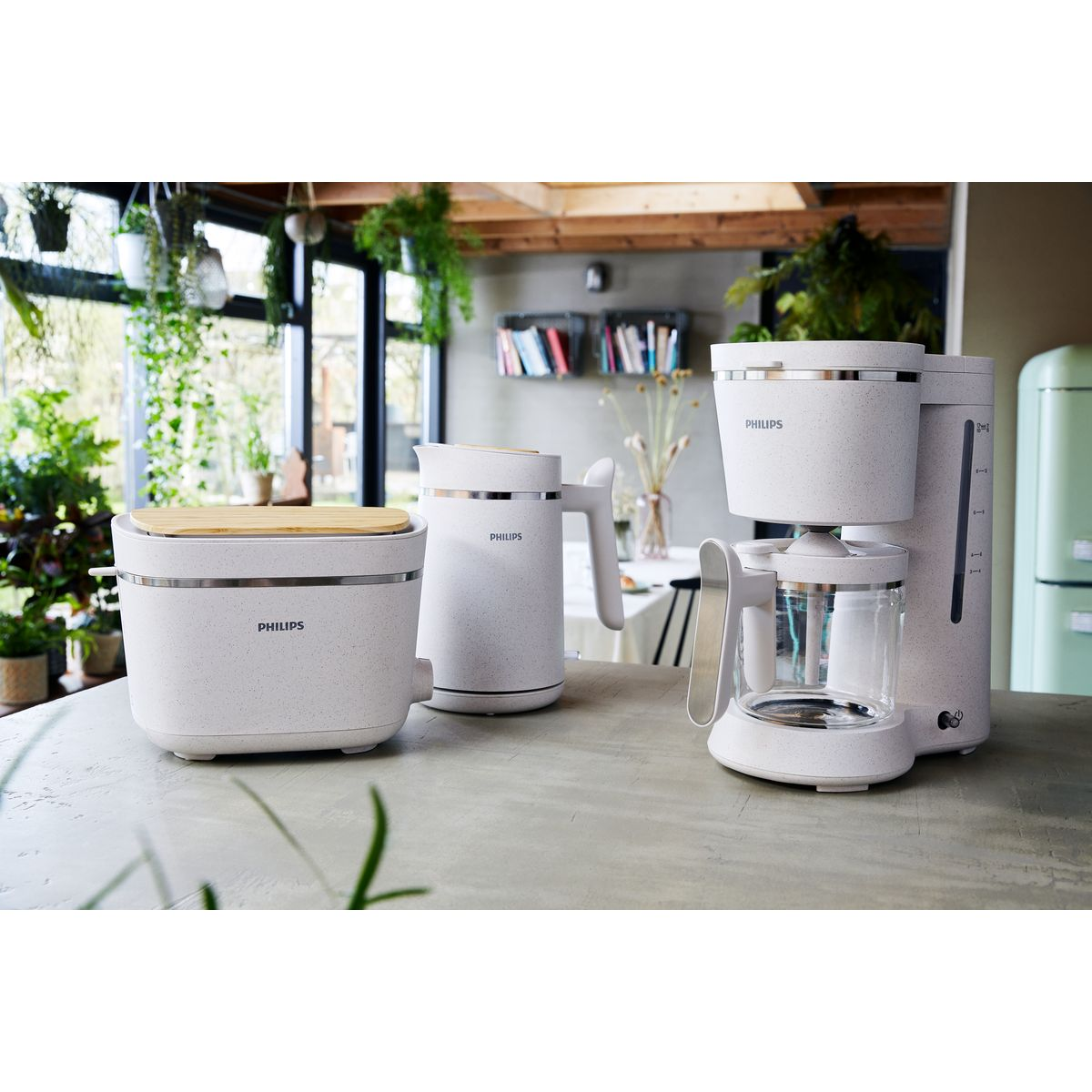 Biały ekspres do kawy, czajnik i toster Philips na stole, z zieloną lodówką i roślinami w tle.