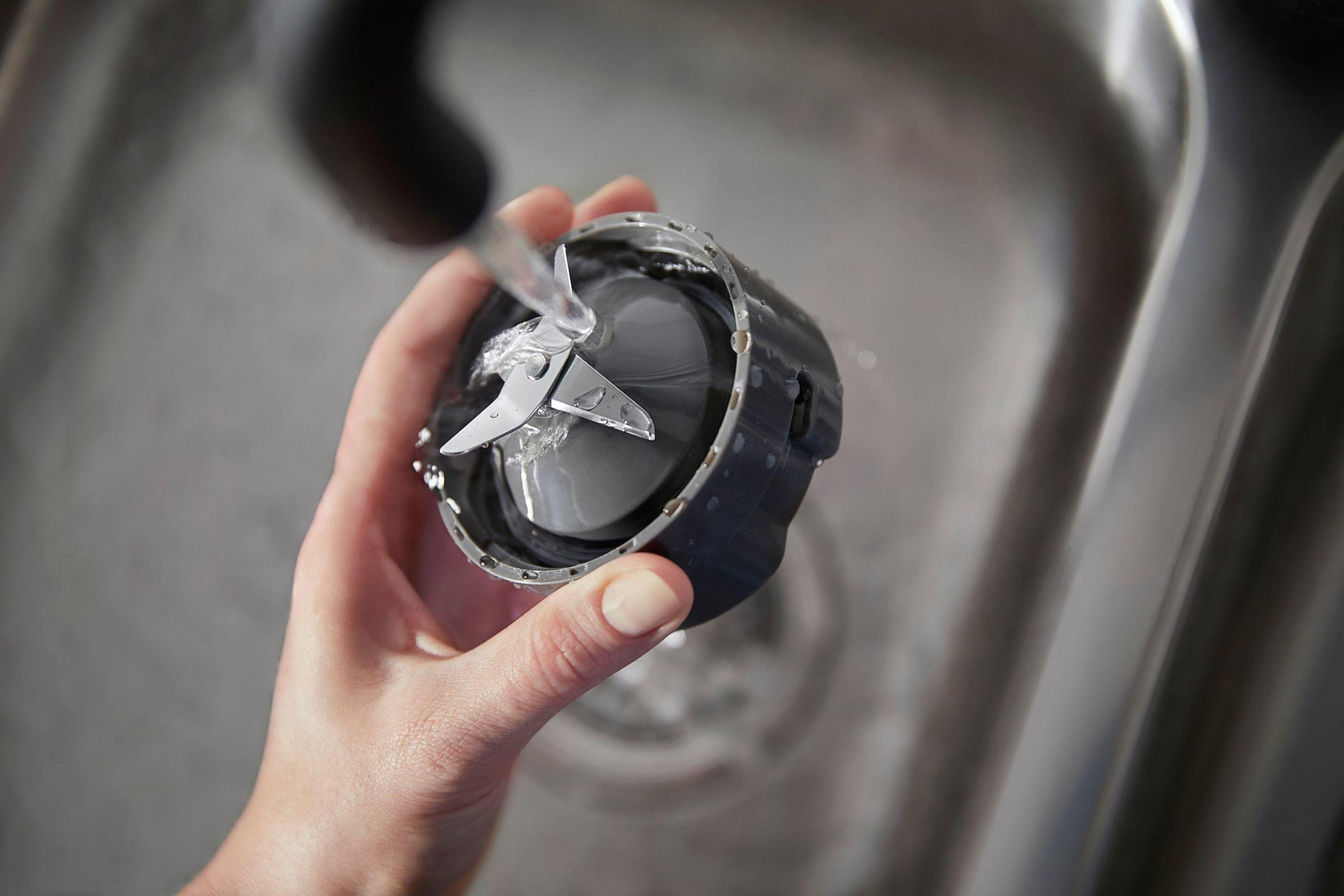 Ręka trzyma część blendera pod bieżącą wodą. Widoczne są srebrne ostrza. Tło to szary zlew.