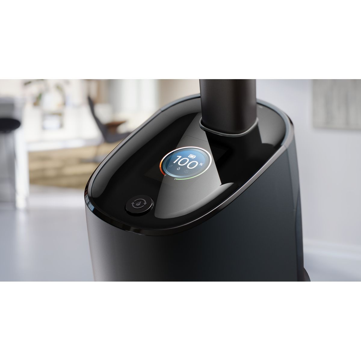 Czarne urządzenie z cyfrowym wyświetlaczem pokazującym 100%. Widoczny jest przycisk zasilania. Tło to rozmyte wnętrze.