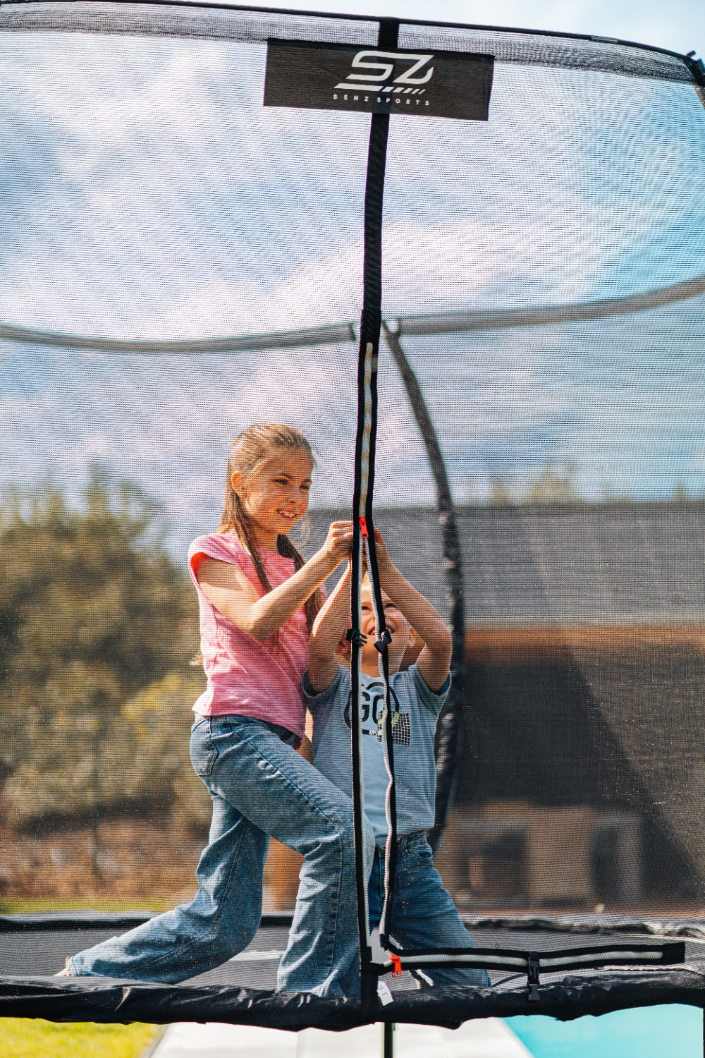 Twee kinderen op een trampoline. Een meisje in roze en een jongen in blauw houden het net vast. De lucht is zichtbaar op de achtergrond.