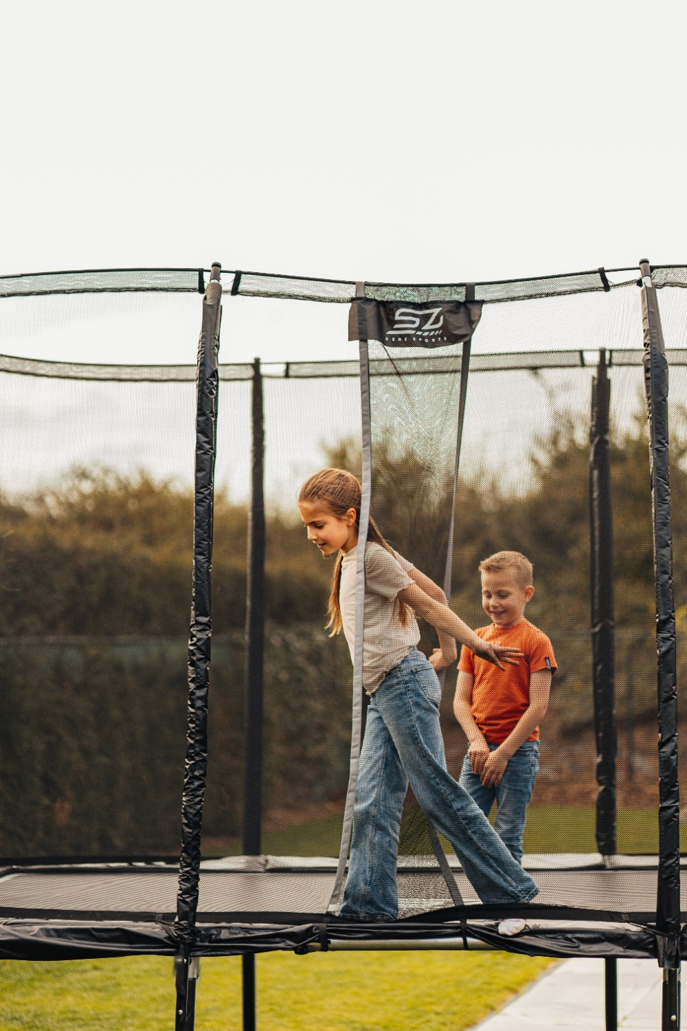 Twee kinderen op een trampoline. Een meisje in een spijkerbroek en een jongen in een oranje shirt spelen lachend op de trampoline.