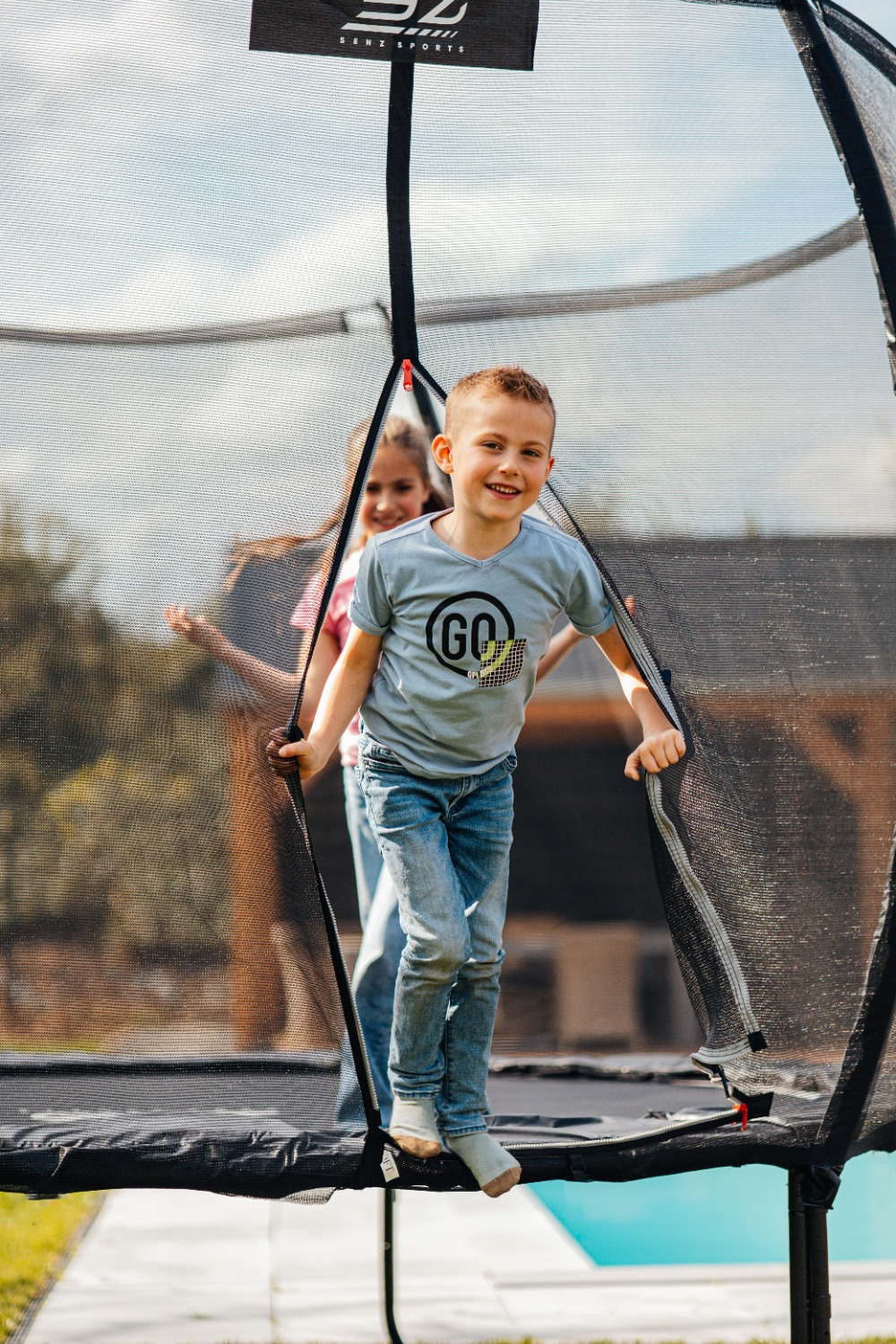 Een jongen glimlacht op een trampoline en houdt het net vast. Er staat een andere persoon achter hem. De lucht is zichtbaar op de achtergrond.