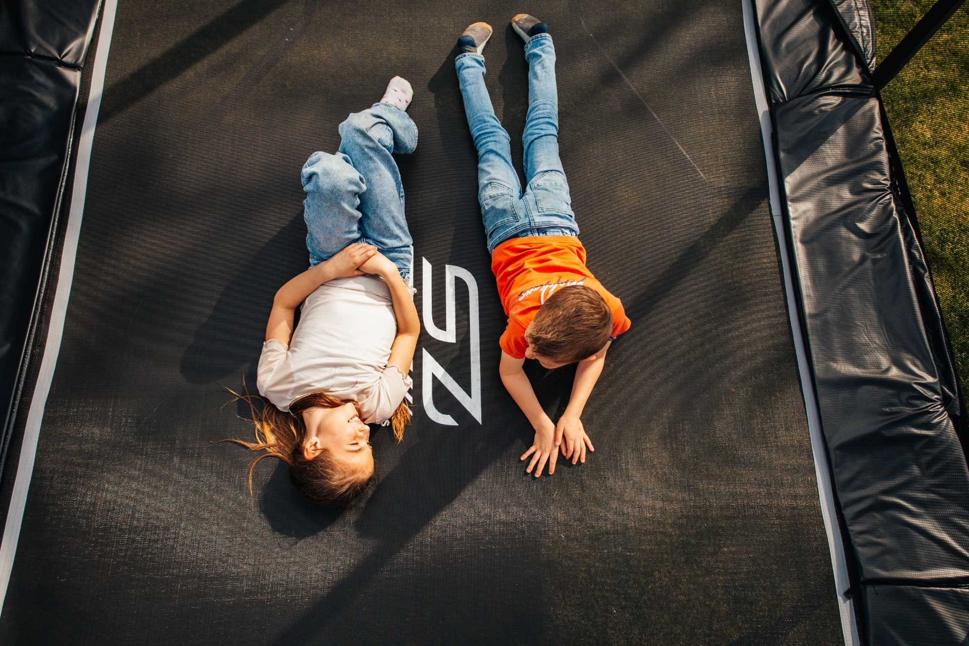 Twee kinderen liggen op een trampoline. De een draagt een wit shirt en een spijkerbroek, de ander een oranje shirt en een spijkerbroek. De trampoline is zwart.