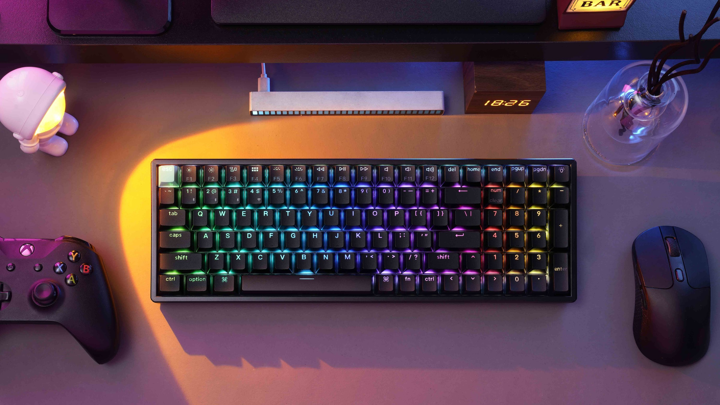 Klawiatura, mysz i gamepad na biurku. Jest też lampa, zegar i wazon z roślinami.