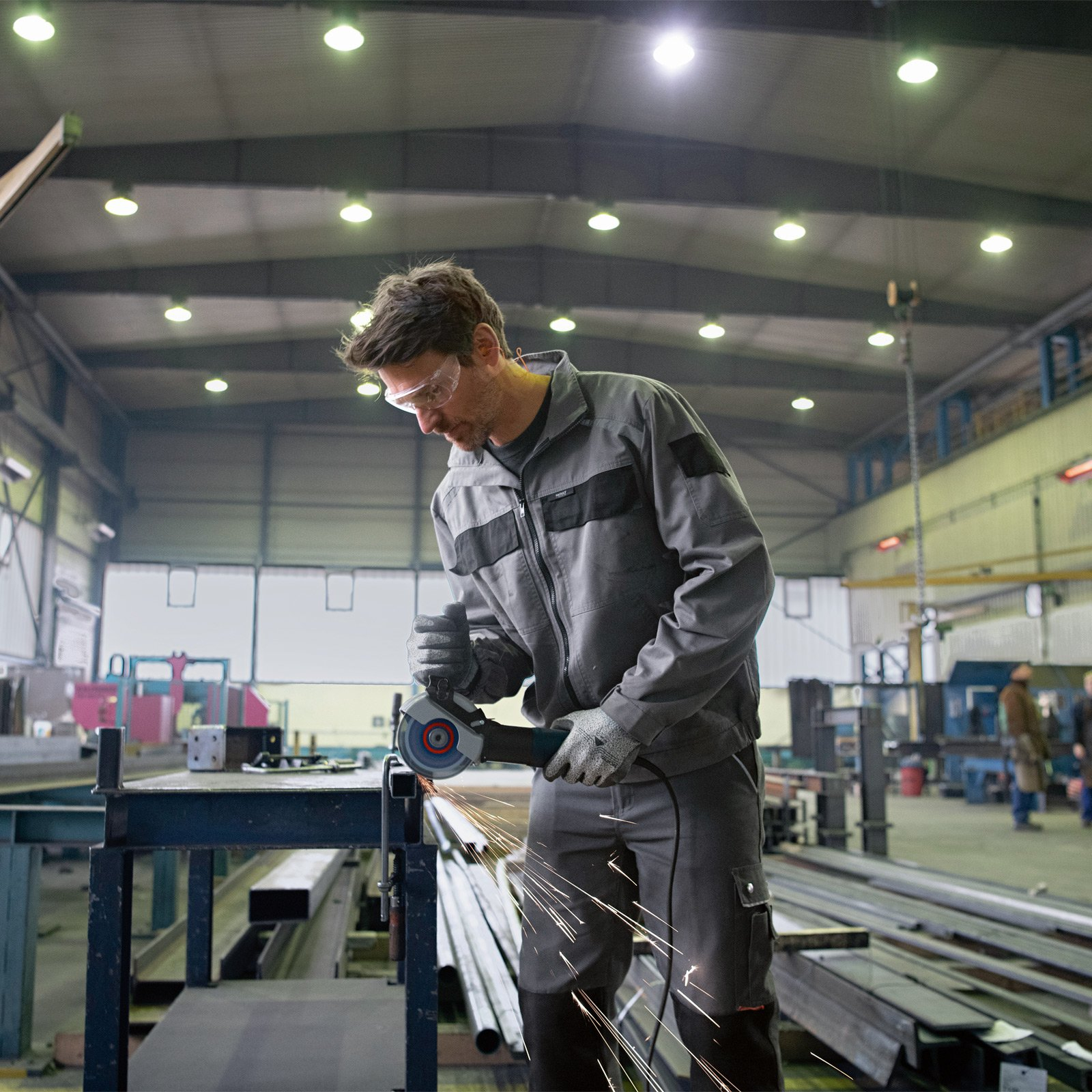 Mężczyzna szlifuje metal szlifierką kątową w warsztacie, lecą iskry. Ma okulary ochronne i odzież roboczą.