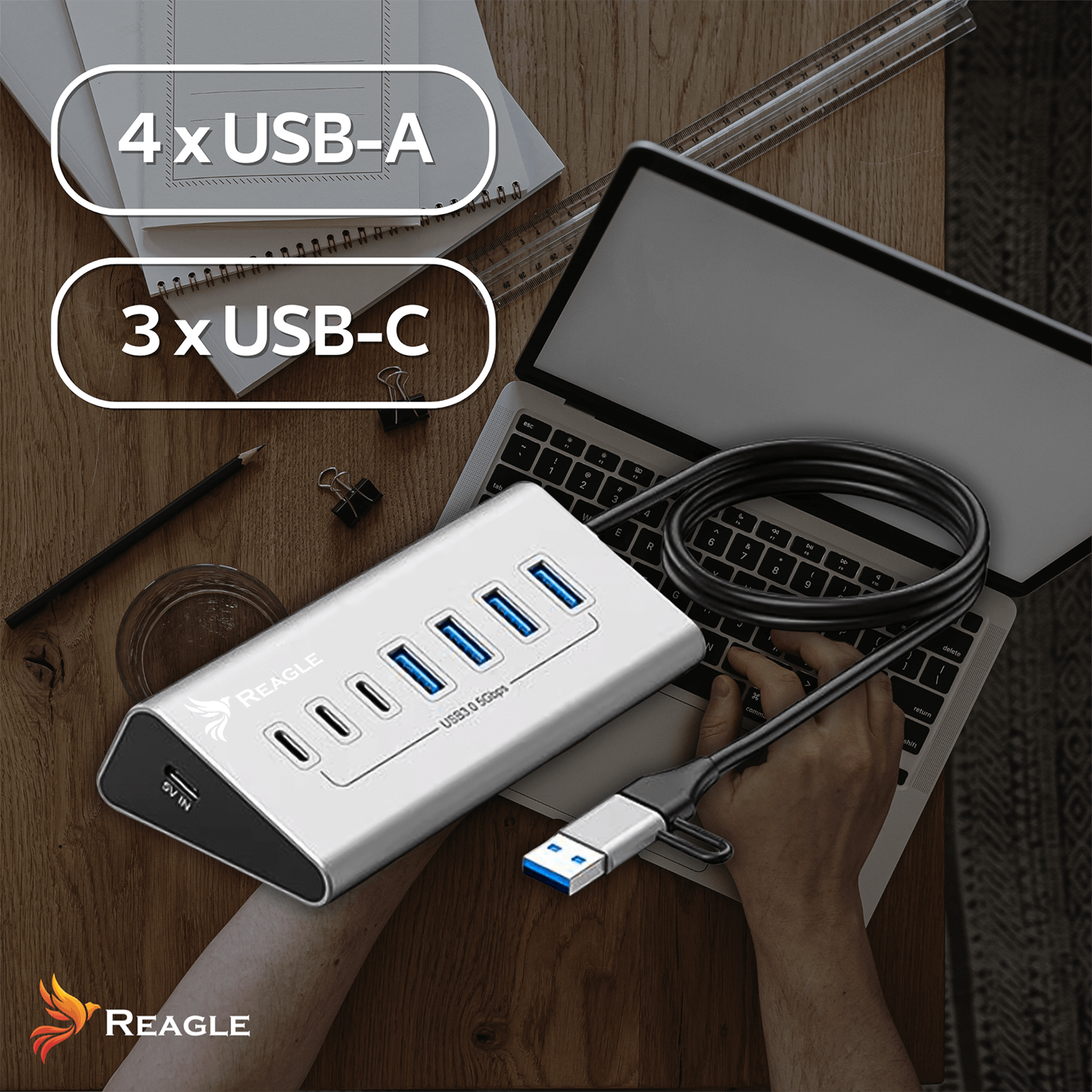 Srebrny hub USB z portami USB-A i USB-C leży na drewnianym stole obok laptopa i linijki. Ręka trzyma kabel USB.