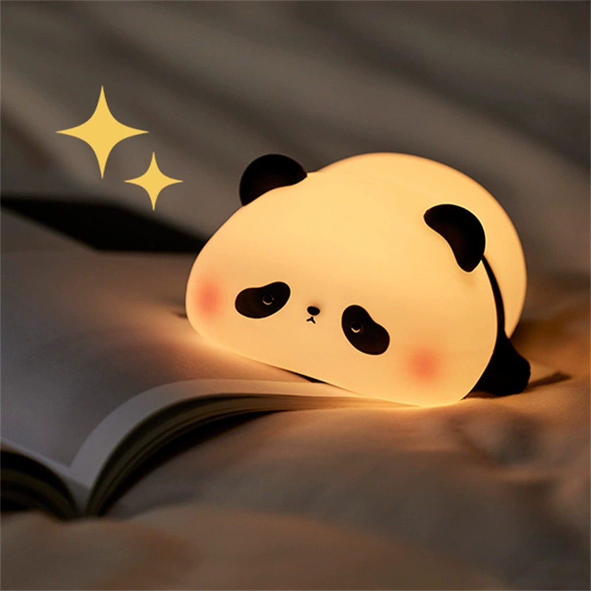 Ein pandaförmiges Nachtlicht liegt auf einem offenen Buch. Es hat schwarze Ohren und Augen, mit rosa Wangen. Zwei gelbe Sterne sind darüber.