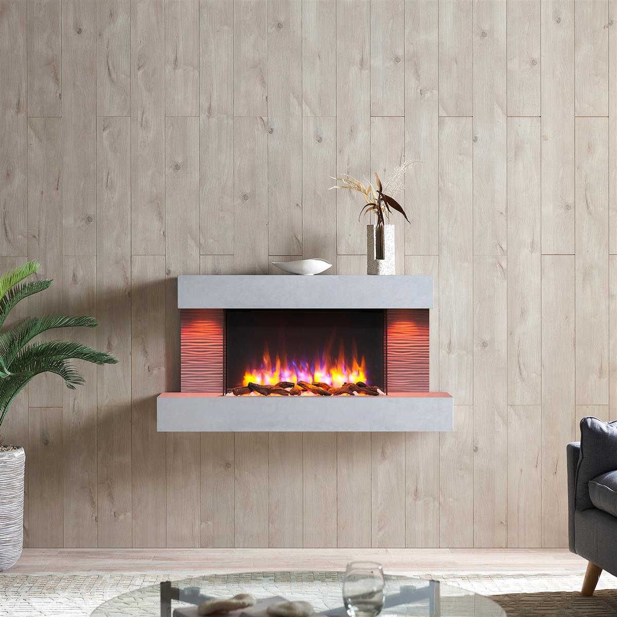 Ein moderner elektrischer Kamin mit Flammen, Holzscheiten und einem grauen Mantel ist an einer hellen Holzwand angebracht. Oben steht eine Vase mit Pflanzen.