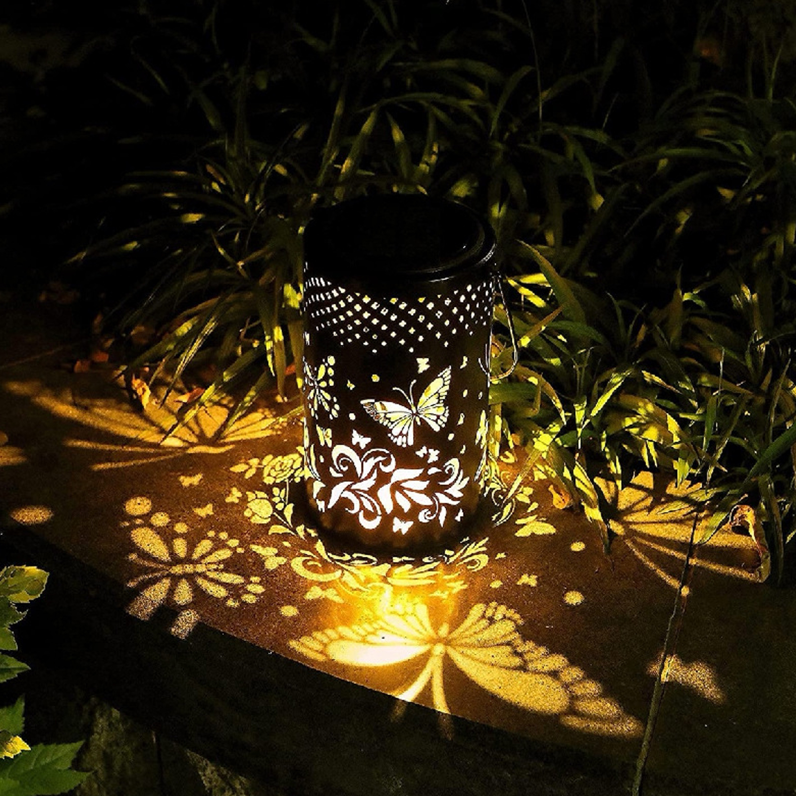 Eine dekorative schwarze Laterne mit Schmetterlings- und Blumenausschnitten wirft ein Lichtmuster auf Stein, umgeben von grünem Laub.