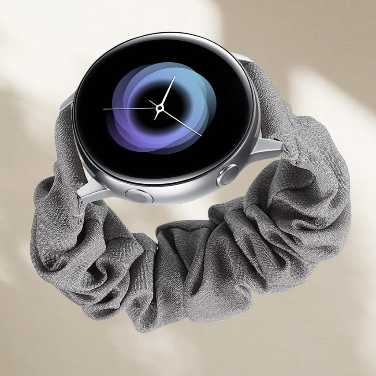 Uno smartwatch argento con schermo nero e lancette bianche, su un elastico grigio. Lo sfondo è una superficie beige chiaro.