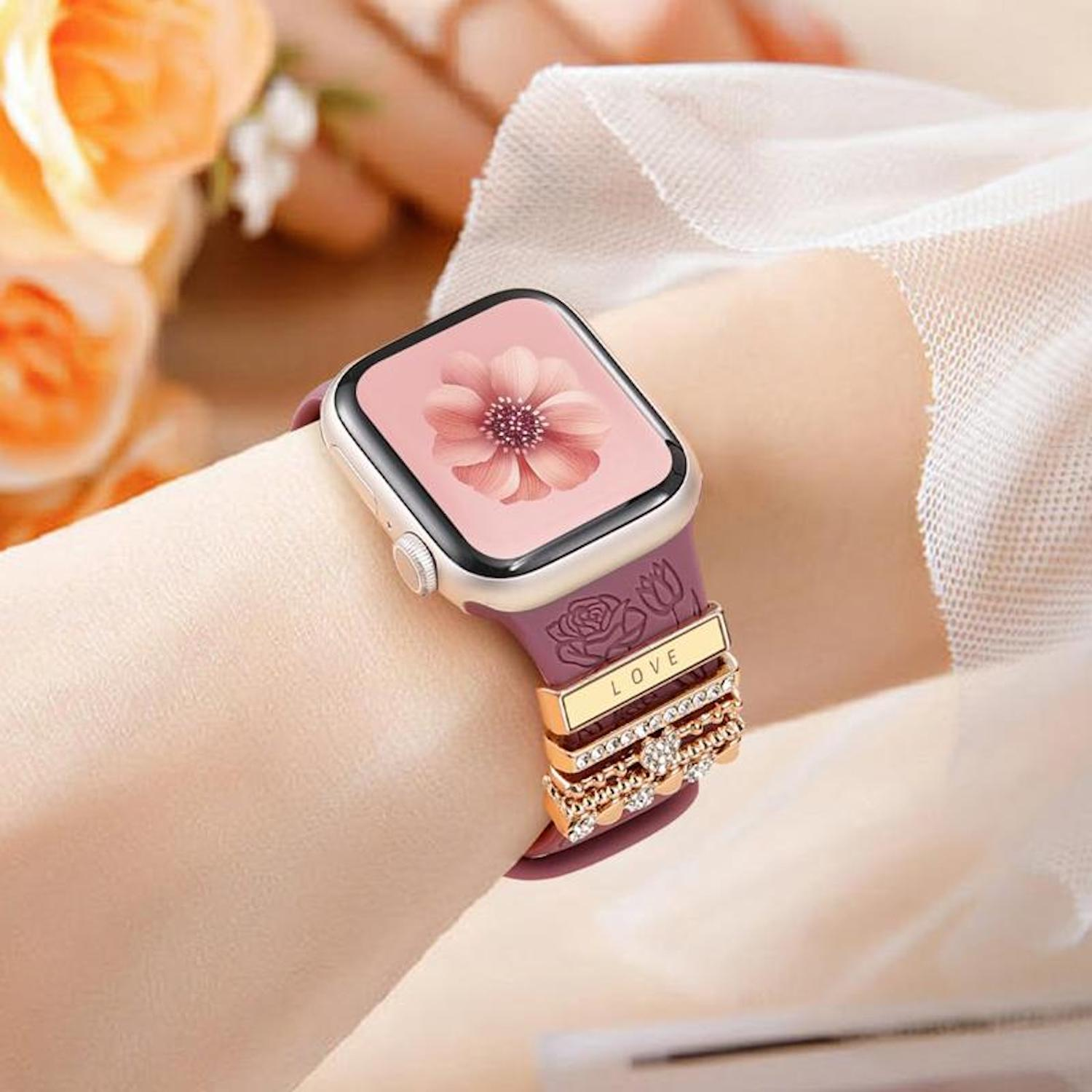 Ein Handgelenk mit einer Smartwatch. Die Uhr hat ein rosa Blumendisplay und ein lila Band mit goldenen Ringen mit der Aufschrift 'LOVE'.