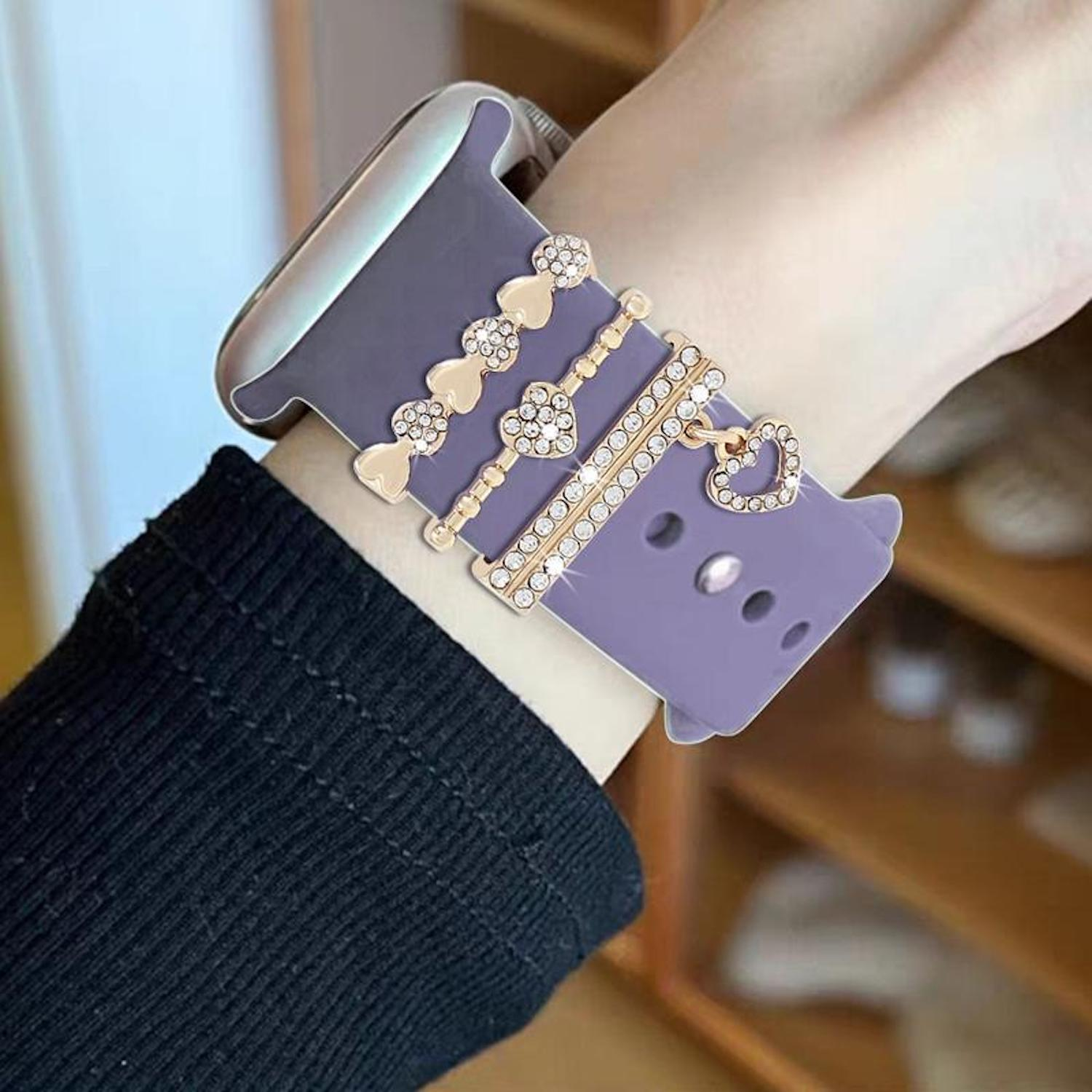 Handgelenk mit lavendelfarbenem Uhrenarmband und goldenen, mit Juwelen besetzten Anhängern. Schwarzer Ärmel sichtbar. Hintergrund zeigt ein Holzregal.