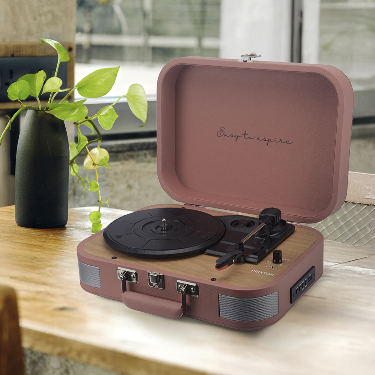 Otwarty gramofon w stylu vintage na drewnianym stole. Po lewej stronie roślina w czarnym wazonie. Tłem jest okno.