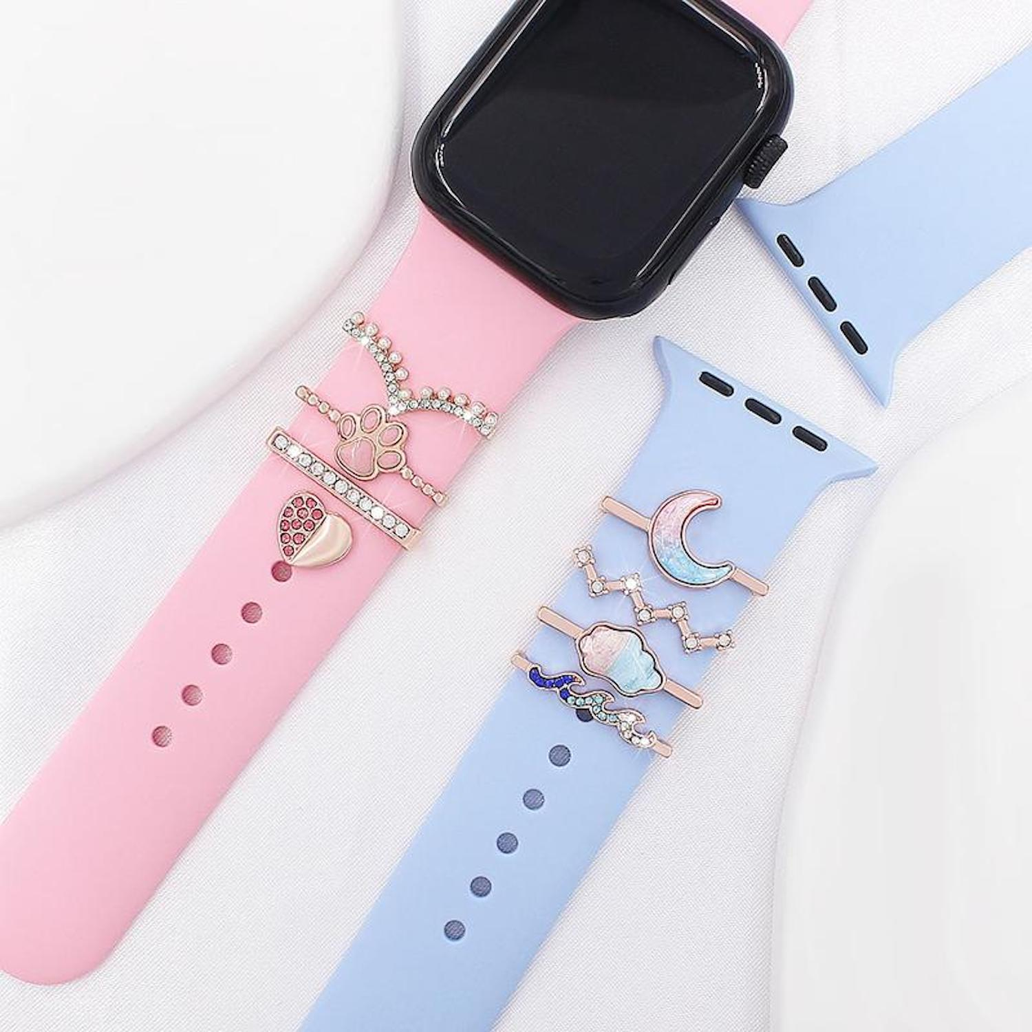 Rosa und blaue Armbänder mit Charms. Eines hat einen Pfotenabdruck, das andere Mond und Wolke. Eine schwarze Uhr ist auf dem rosa Band.