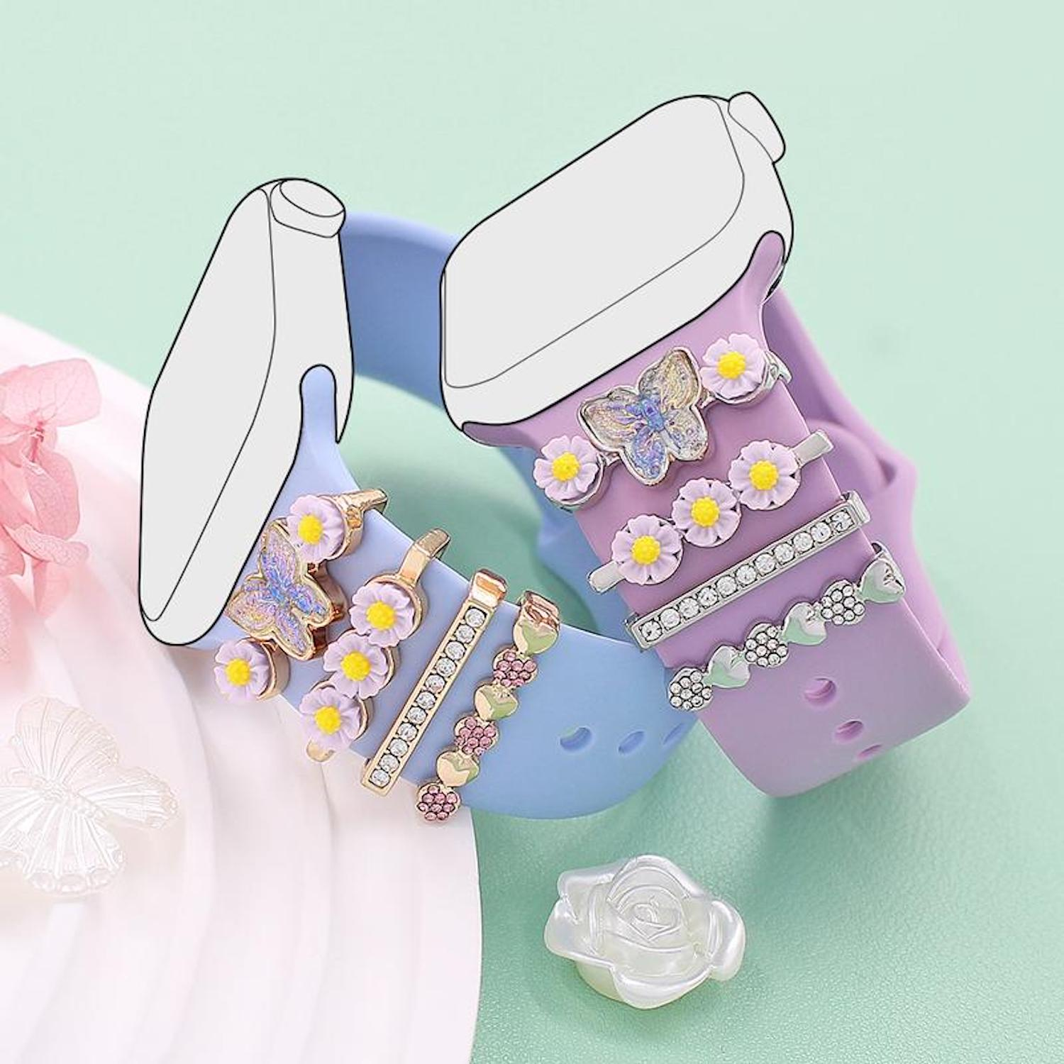 Das Bild zeigt zwei Smartwatch-Armbänder, lila und blau, mit dekorativen Charms, darunter Schmetterlinge, Blumen und ein Herz. Unten ist eine weiße Rose.