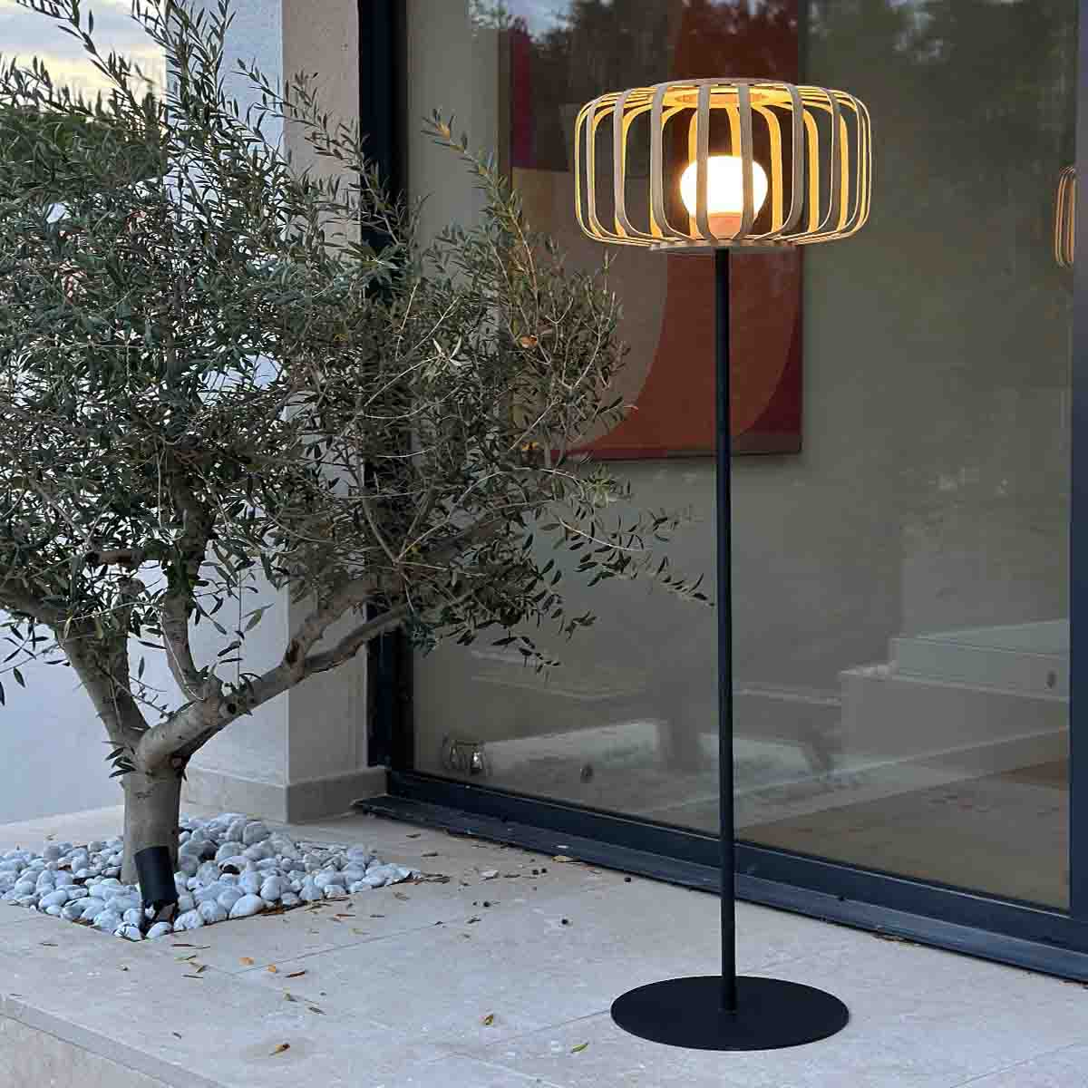 Eine Außenszene zeigt eine schwarze Lampe mit rundem Schirm. Links ein Olivenbaum. Rechts eine Glastür.