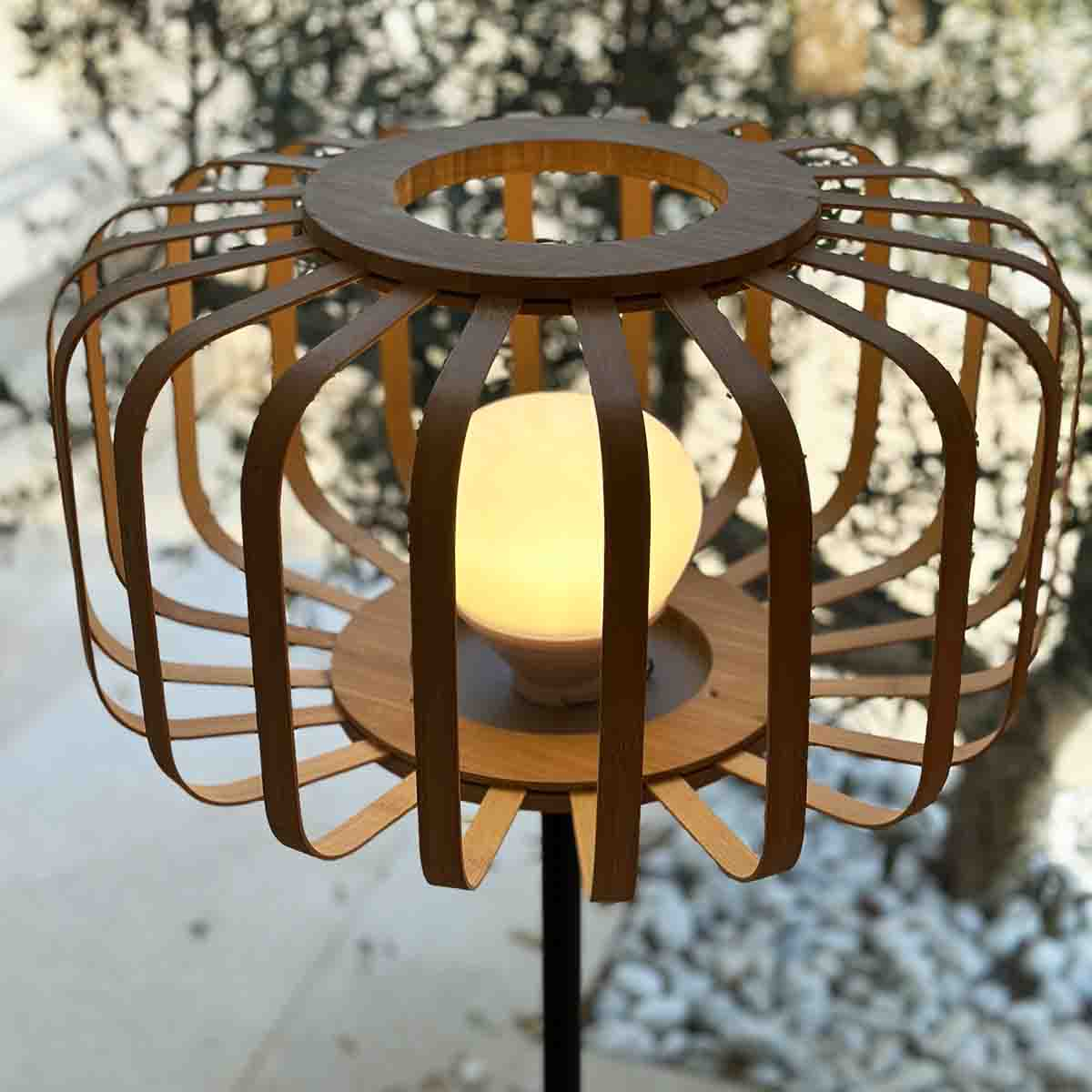 Eine Holzlaterne mit leuchtender Glühbirne. Sie hat ein käfigartiges Design. Hintergrund: Bäume und weiße Steine.